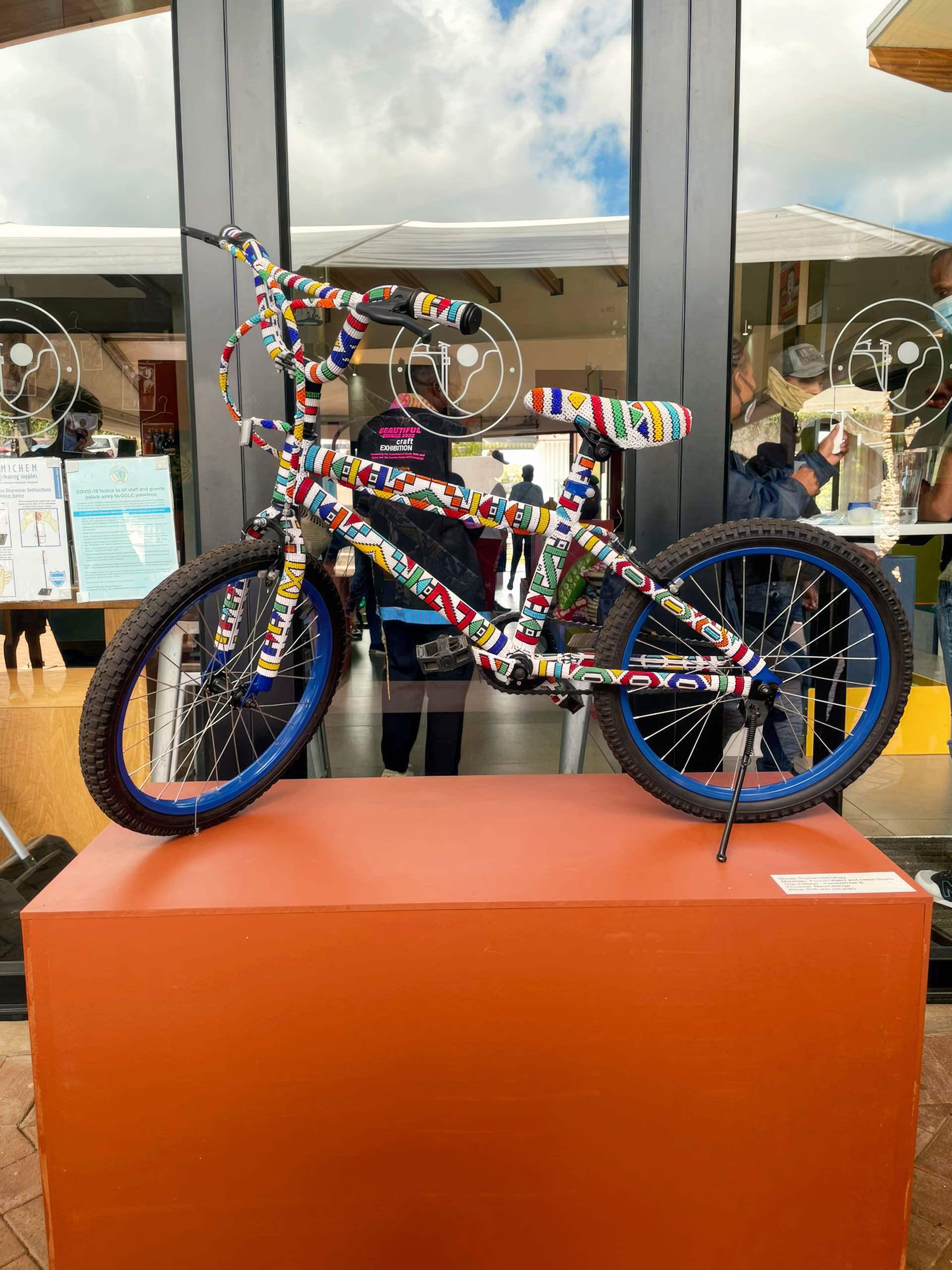 A BMX bicycle covered in colourful beadwork sits on an orange plinth in front of glass doors. The intricate bead patterns are geometric and vibrant, wrapping the entire frame, handlebars, and fork. Reflections and people are visible in the background through the glass.