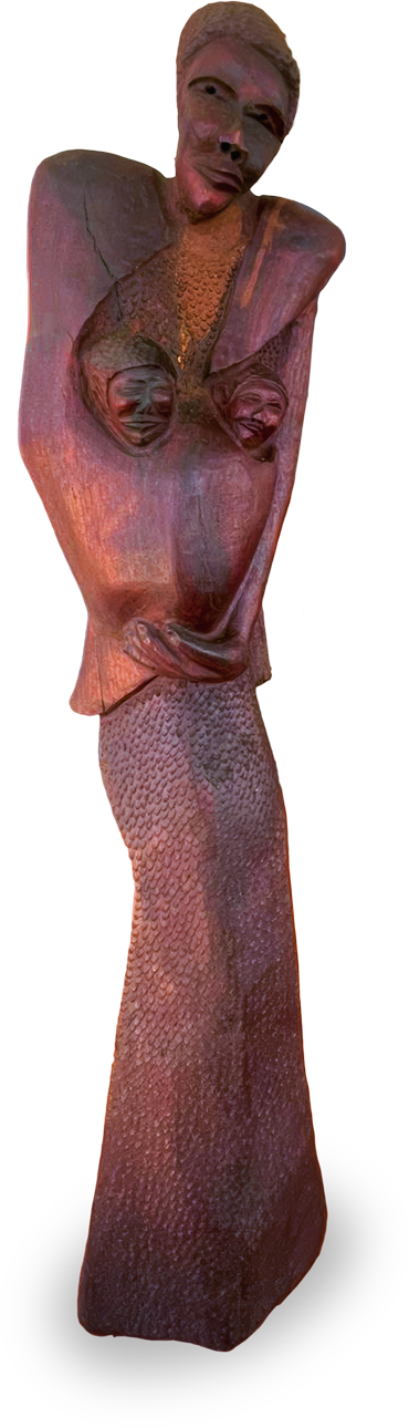 A tall, textured wooden sculpture of a woman with exaggerated, angular shoulders, prominent breasts, and arms crossed below her chest. She wears a long, close-fitting skirt, and her facial features are carved with a serene, closed-mouth expression.