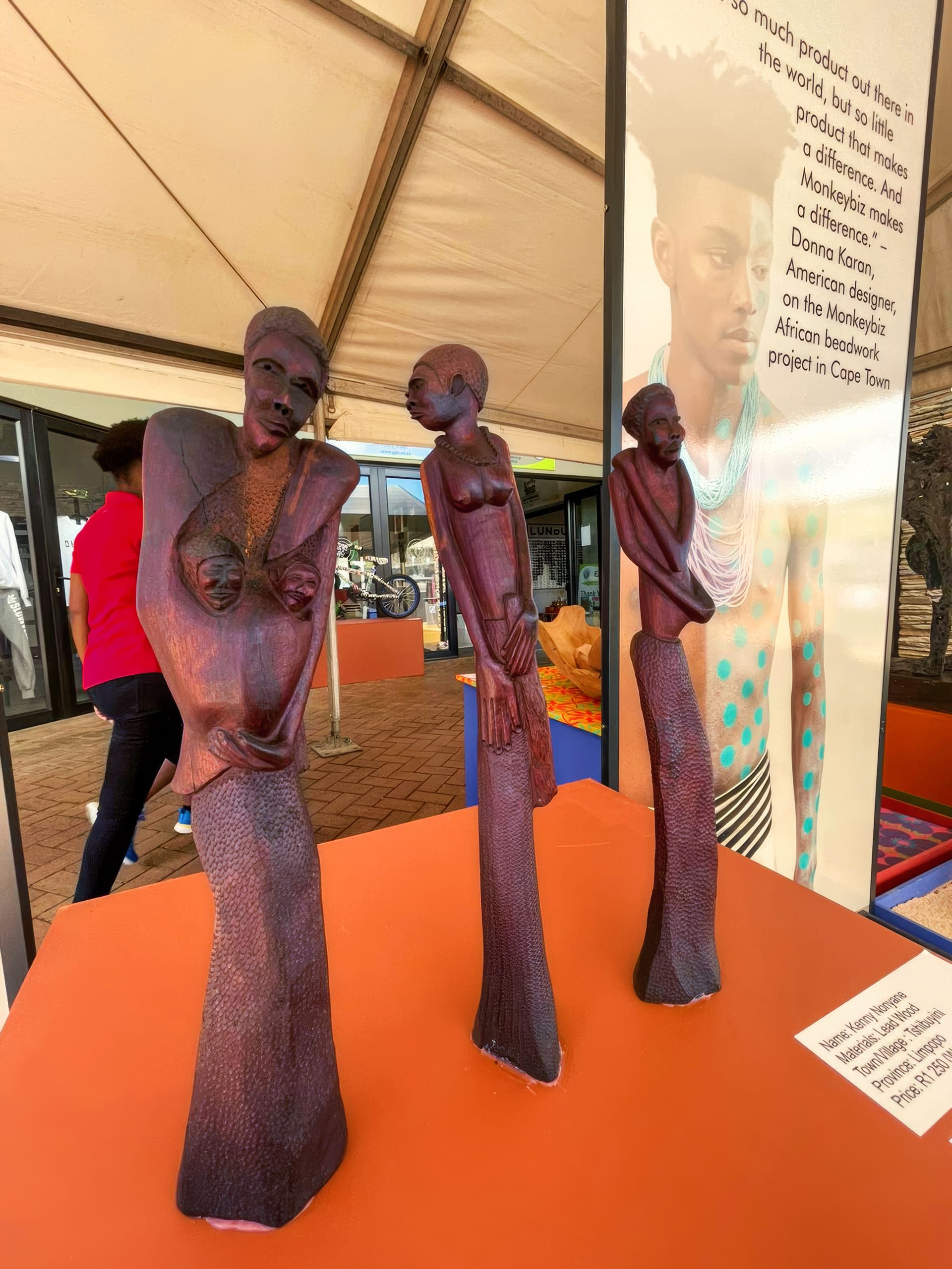 Three tall, slender wooden sculptures of stylised women stand on an orange table. Each figure has elongated features and textured dresses. Behind them, a poster shows a portrait of a person with styled hair and jewellery, set under a tent with people in the background.