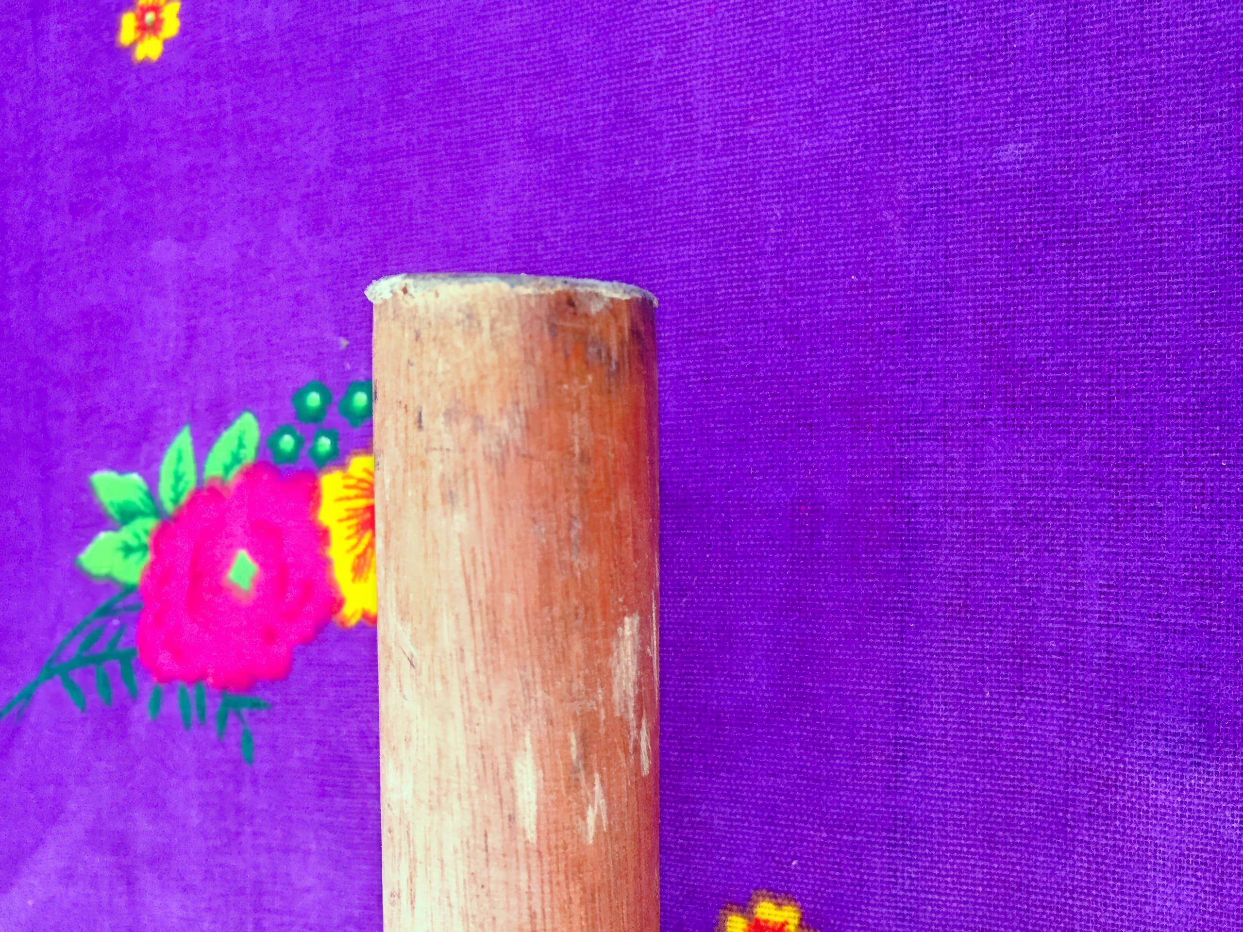 A wooden cylindrical stick stands upright on a purple fabric background, decorated with bright red, yellow, and green floral designs on the left side. The stick appears weathered and slightly rough.