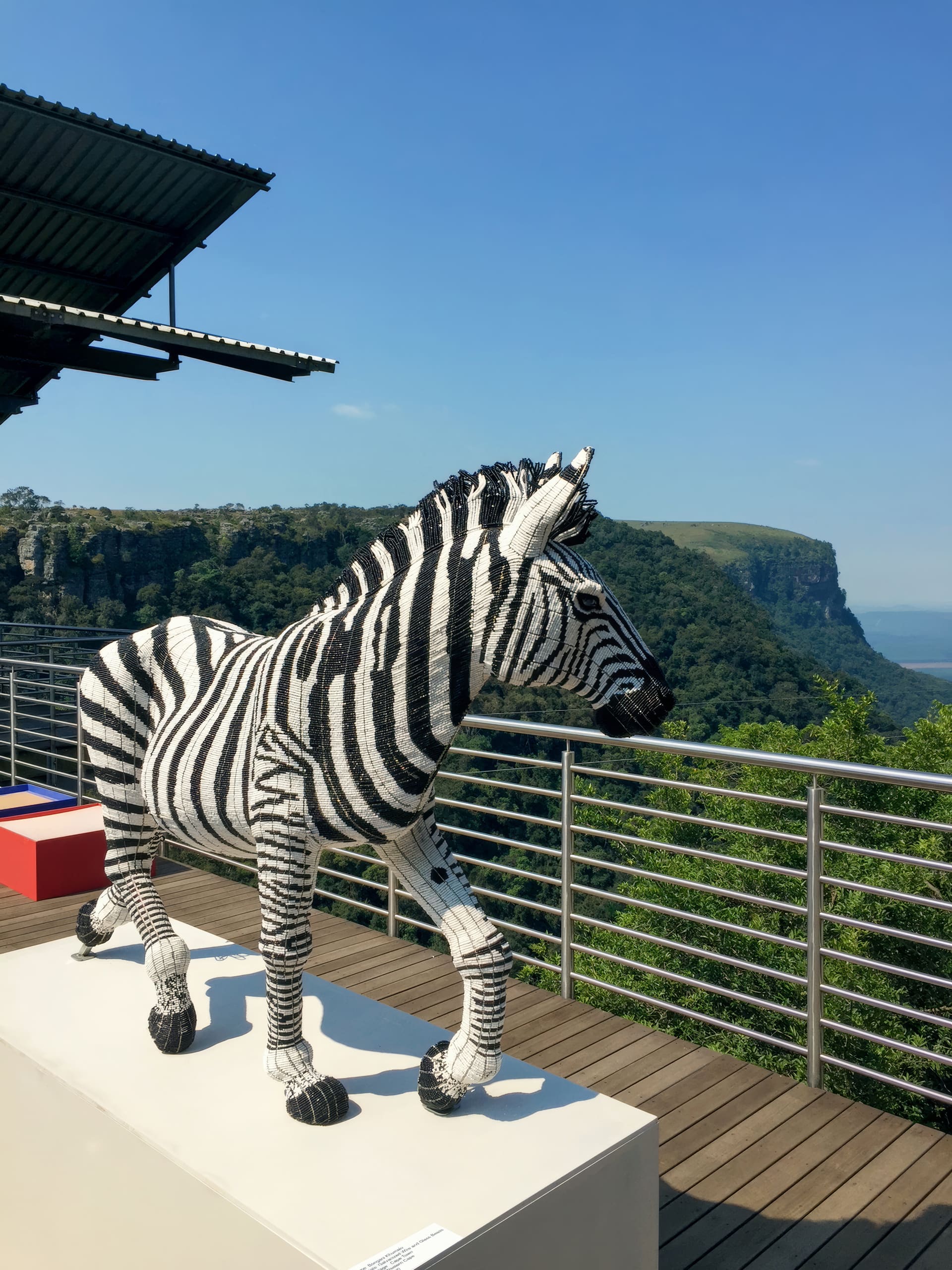 A life-sized zebra sculpture made from LEGO bricks stands on a white plinth on a wooden decking with a metal railing. Green forested cliffs and a blue sky form the scenic backdrop. The area is partially shaded by a metal roof.