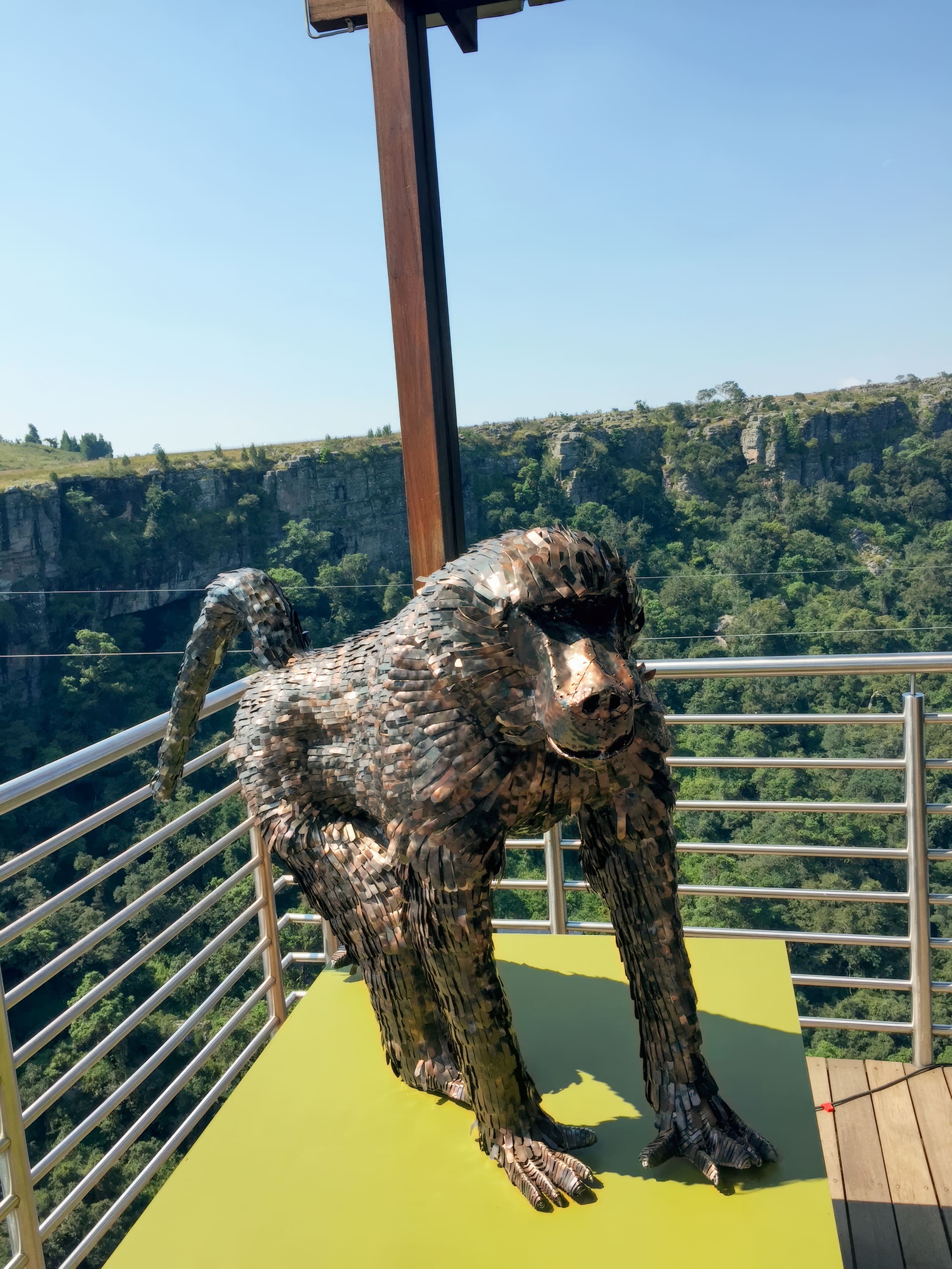 A metallic baboon sculpture stands on a yellow platform, enclosed by a silver railing. Behind it, there is a lush, green forested gorge and cliffs under a clear blue sky, suggesting a scenic viewpoint.