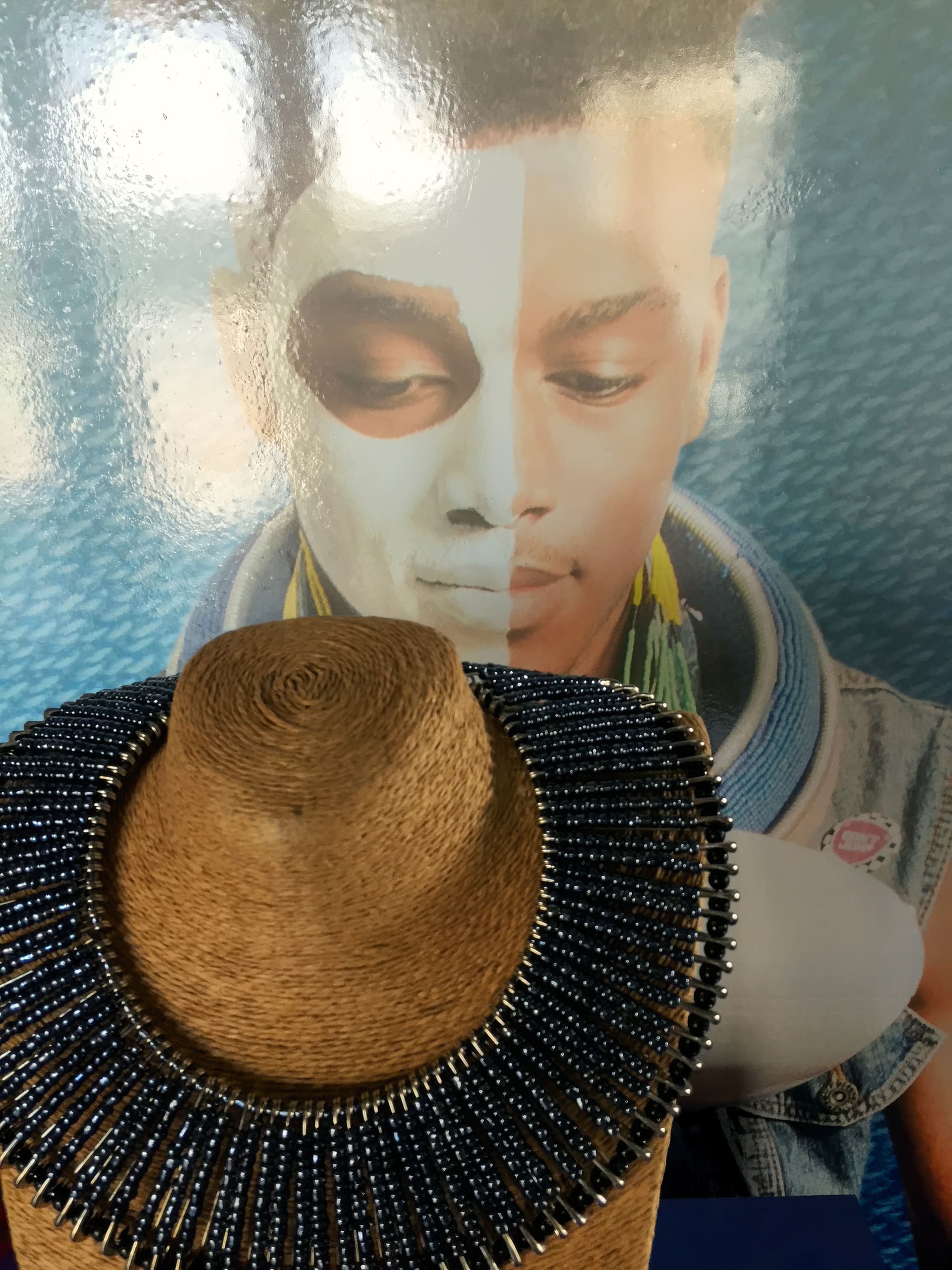 A beaded necklace displayed on a woven stand is in the foreground. Behind it is a poster of a young man with half of his face painted white, the other half bare, and he is wearing colourful earrings and a denim jacket, against a blue background.