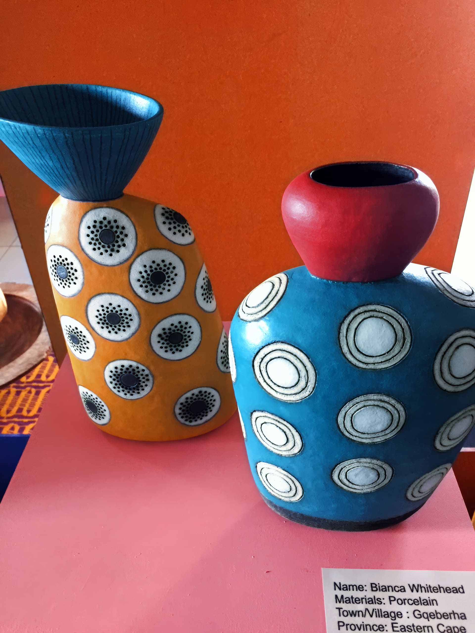 Two porcelain vases with bold geometric designs sit on a pink surface. The left vase is yellow with black and white dot circles; the right is blue with white rings and a red rim. An orange wall is behind them, and a label with artist details is partially visible.