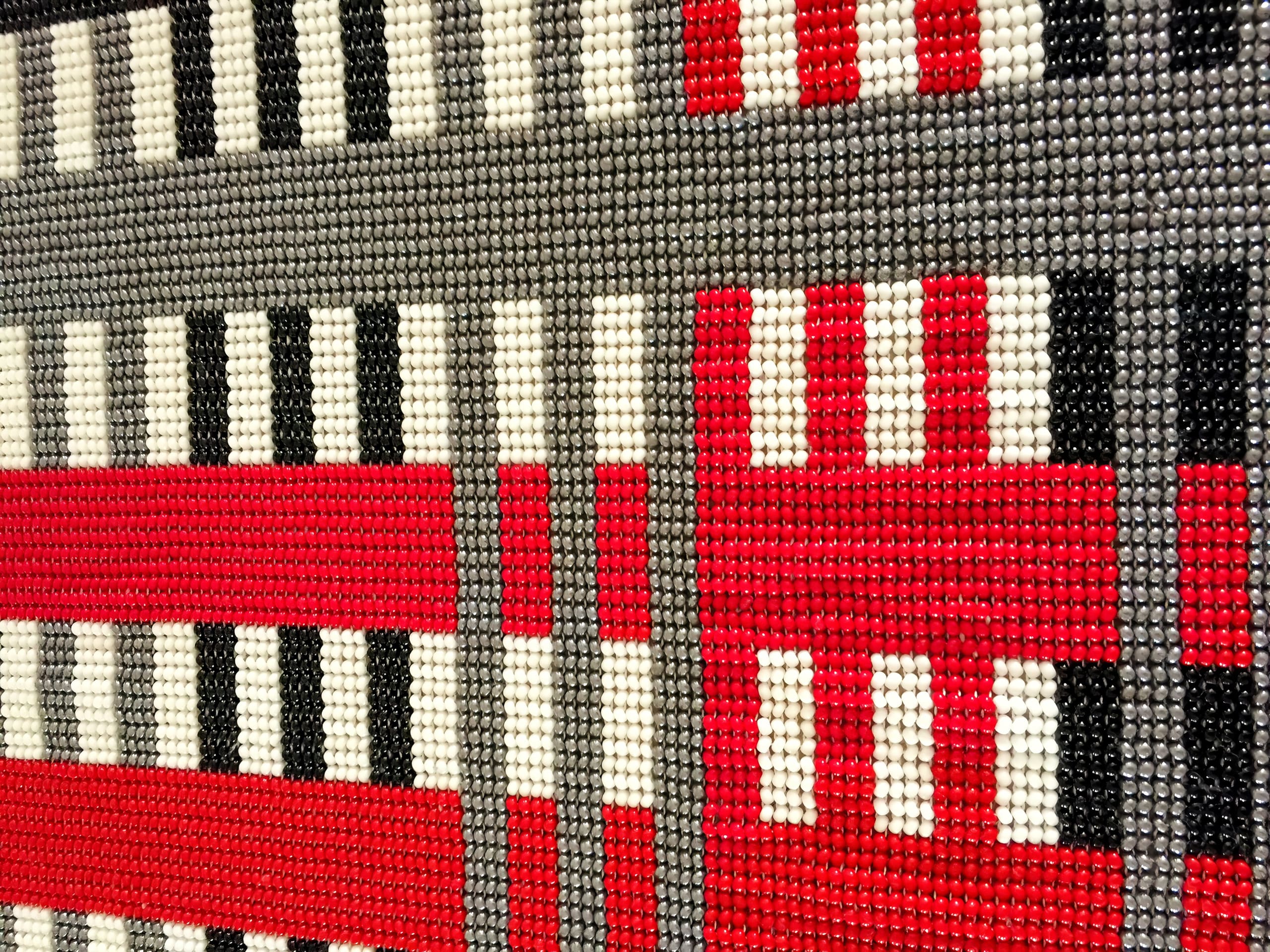 A close-up of a grid pattern made from small, round beads in red, white, black, and grey. The design features alternating horizontal and vertical stripes, creating a bold, geometric, check-like abstract composition.
