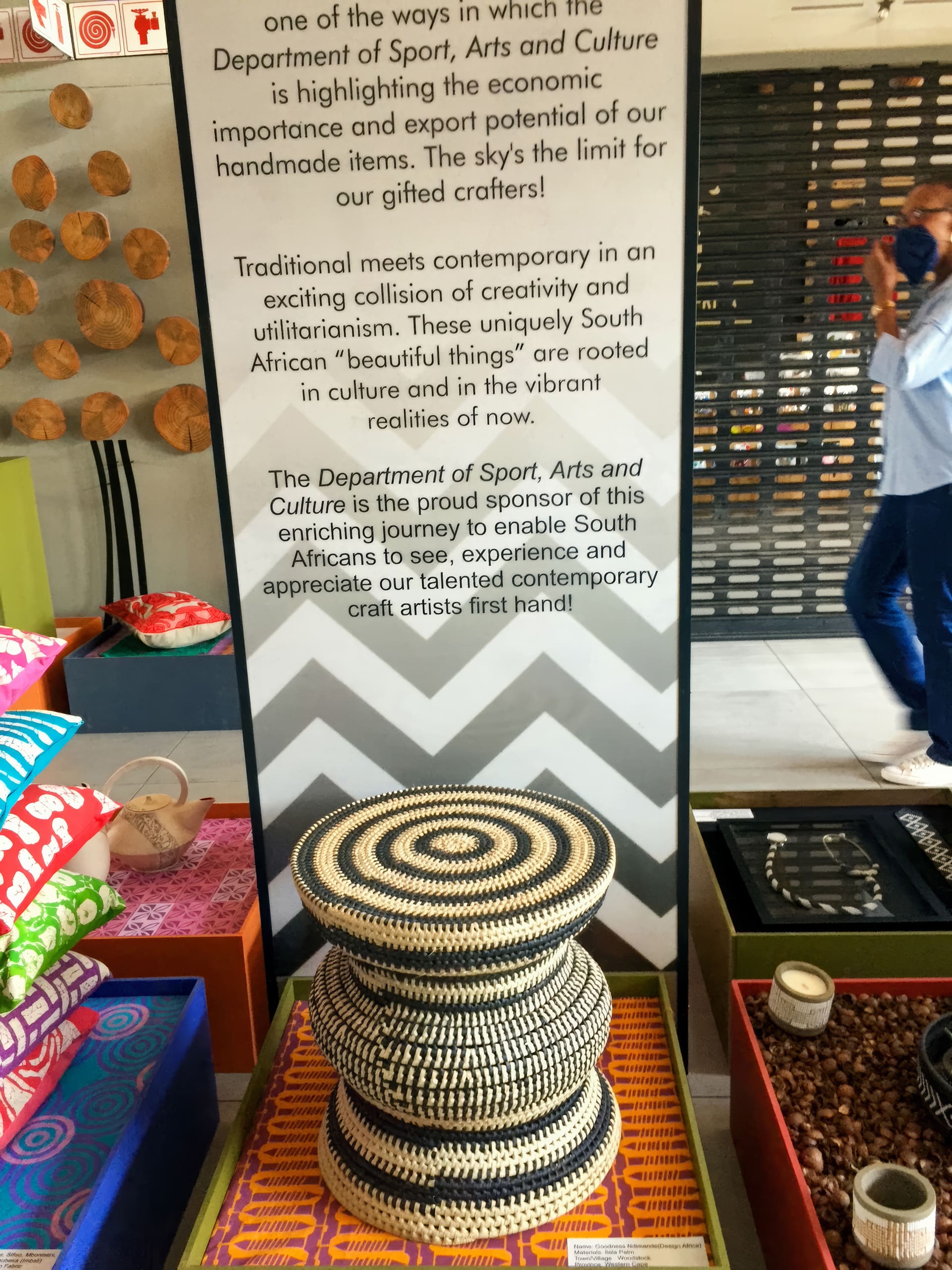 A display at an exhibition features a sign about South African craft, supported by the Department of Sport, Arts and Culture. In front, two woven black-and-white baskets are stacked. Bright patterned textiles and wooden crafts surround the display.