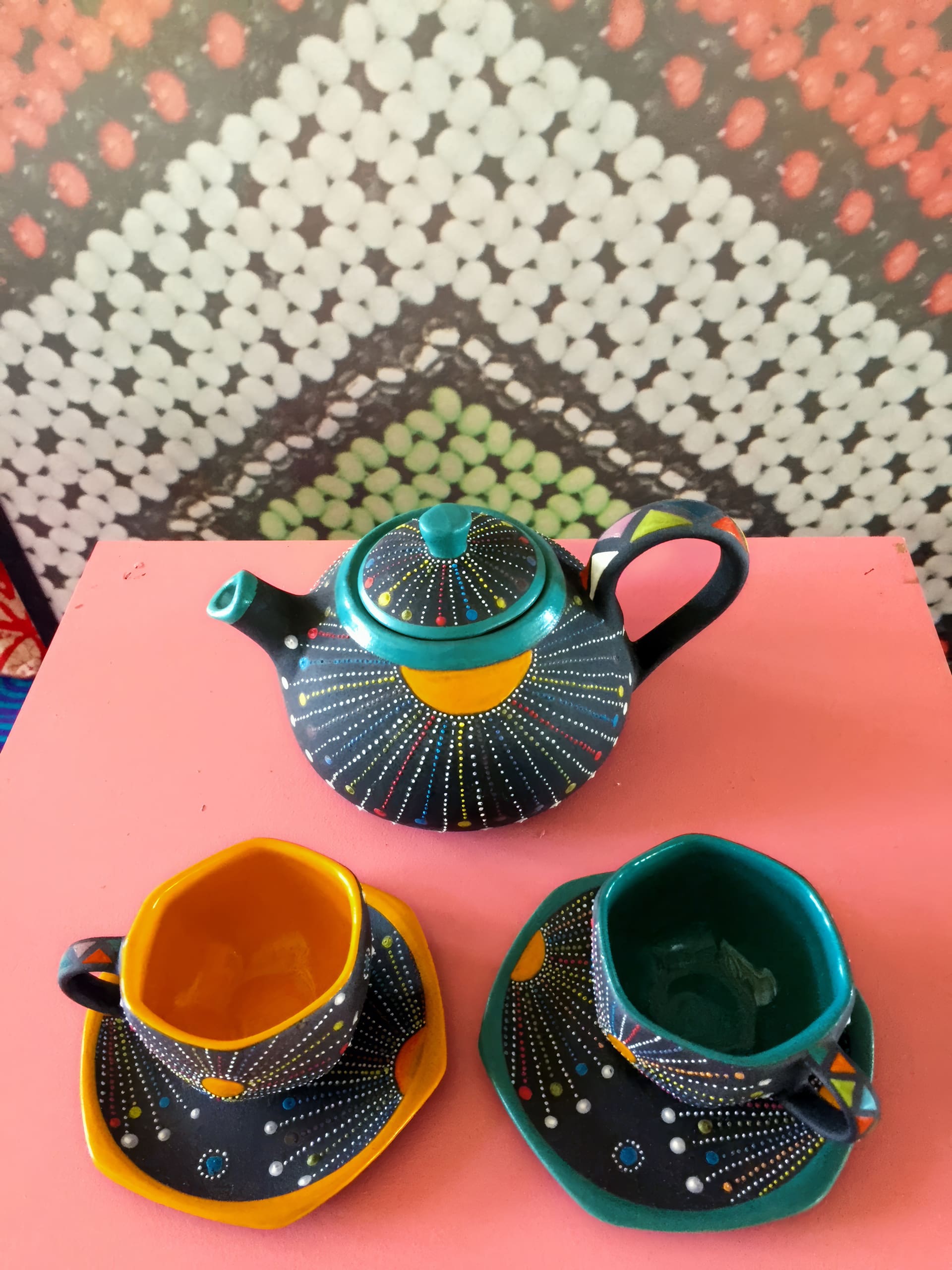 A colourful ceramic tea set sits on a pink surface. The set includes a teapot and two cups with matching saucers, all decorated with bright dots and geometric patterns. The cups' interiors are yellow and teal, and a patterned backdrop is visible behind them.