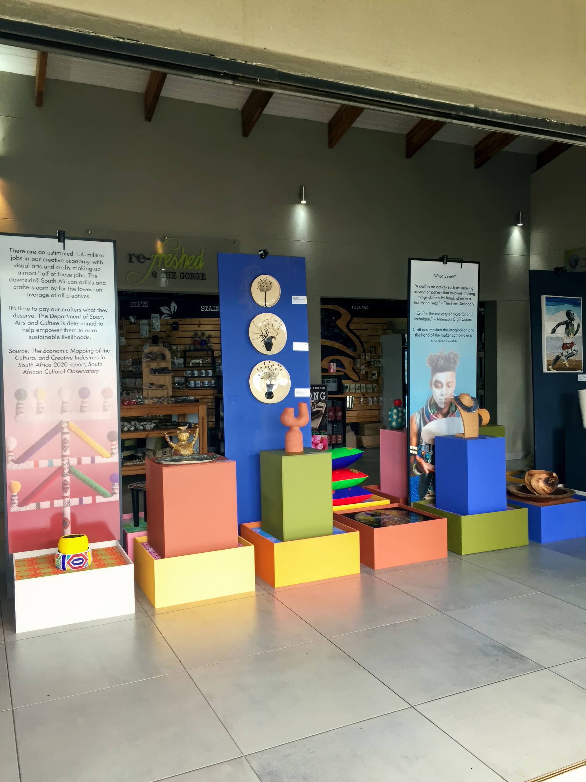 A museum-style display with colourful plinths holds various art pieces and pottery. Large vertical panels with text and images stand behind, providing exhibition information. The backdrop includes shelves of products and a wooden ceiling with exposed beams.