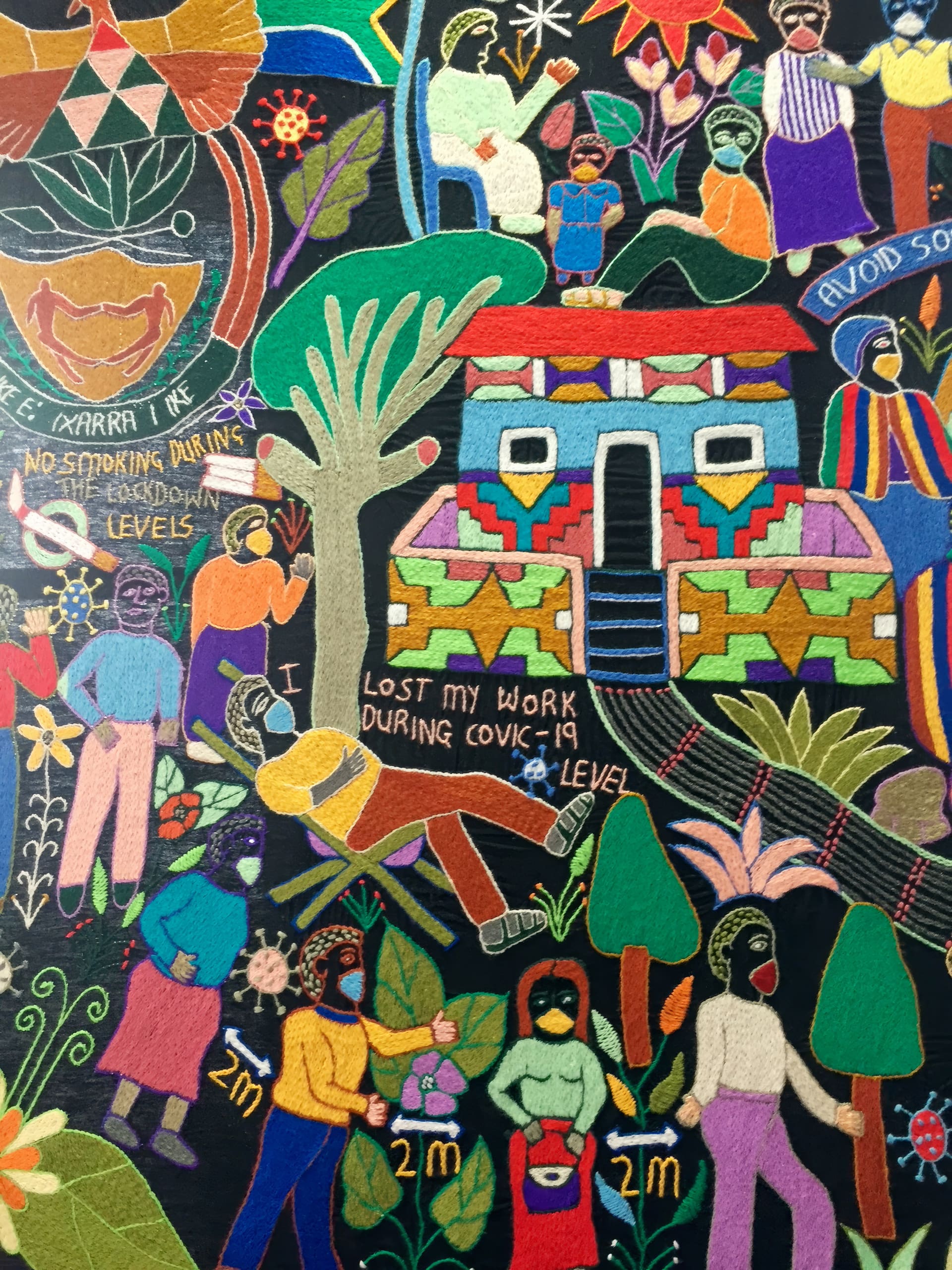 Colourful embroidered artwork depicts people in masks, standing 2 metres apart. Text reads “I lost my work during COVID-19 lockdown levels.” Other stitched messages urge social distancing and no smoking. There are stylised plants, houses, and a rainbow in the background.