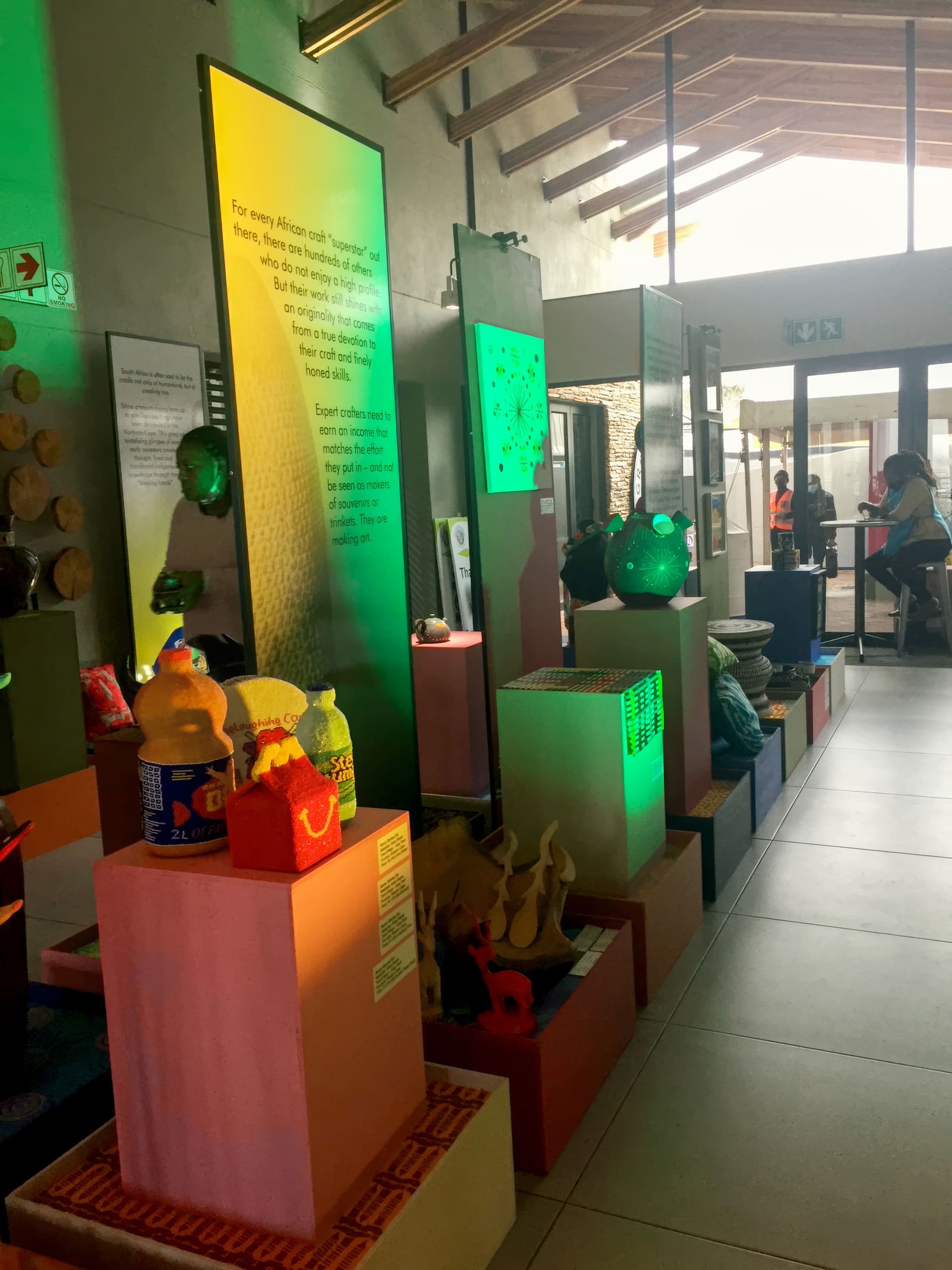 A museum exhibition with colourful displays on plinths, featuring bottles, sculptures, and crafts. Large illuminated panels with text are in the background. Natural light enters from glass doors, and a person stands near the entrance. The setting is modern and well-lit.