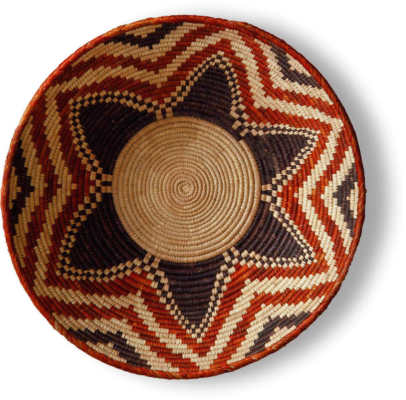 A woven basket with a circular shape, featuring a starburst pattern. The design alternates between dark brown, orange-red, and cream zigzag sections radiating from the centre, with concentric rings at the basket’s round base. The background is black.