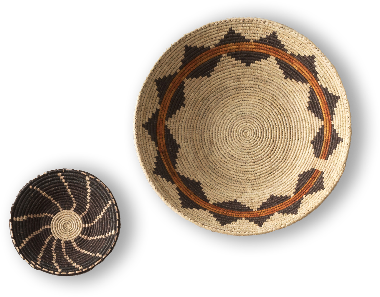 Two woven baskets are displayed on a black background. The larger basket has a tan base with brown and black geometric patterns around the rim. The smaller basket is darker, featuring a black base with a spiralling tan pattern radiating from the centre.
