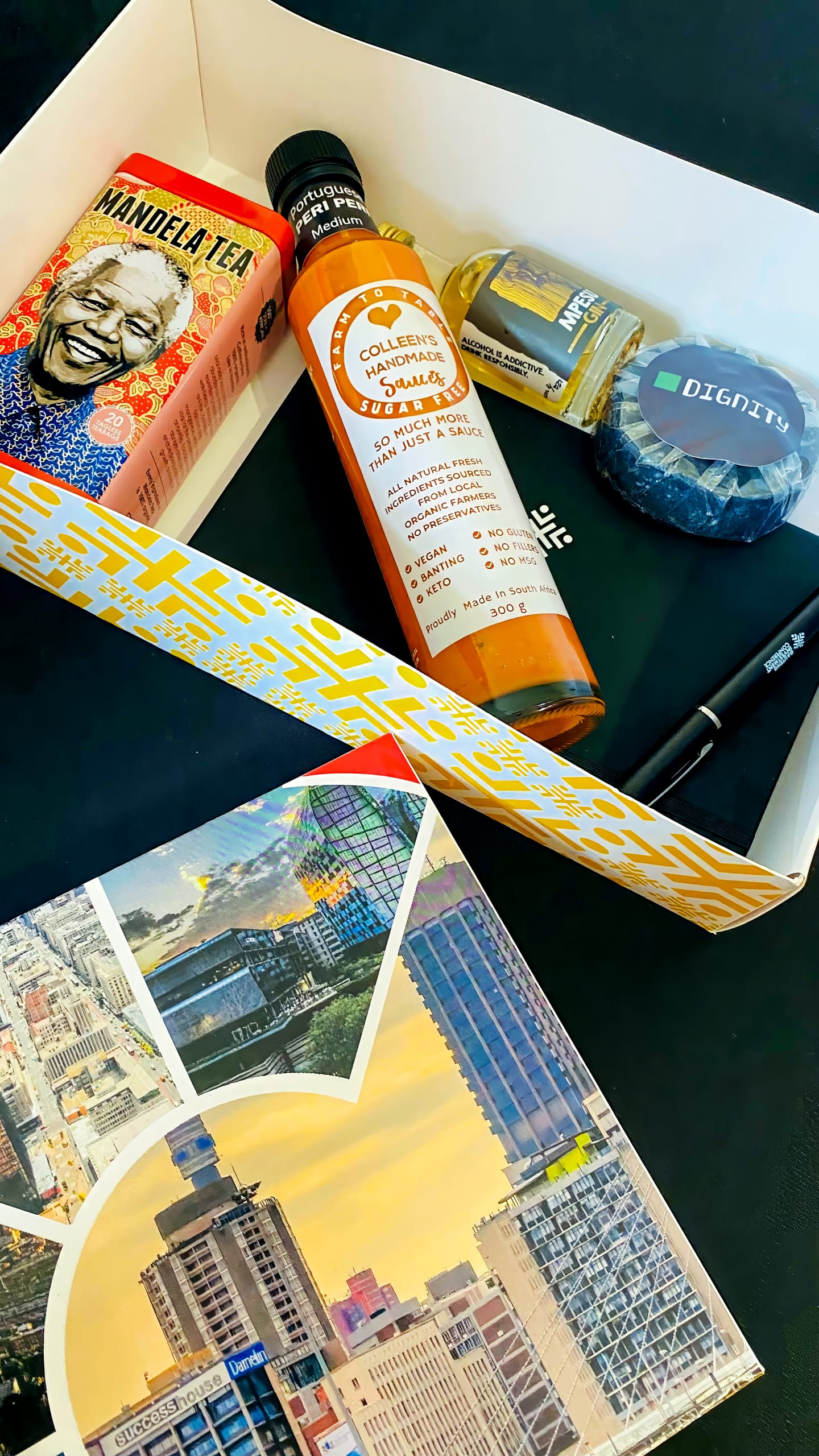 A gift box contains Mandela Tea, a bottle of Colleen's Homemade Hot Sauce, a Dignity branded item, a box of Mentos, a black pen, and a postcard featuring cityscape photos. The box’s lid shows a bold, yellow geometric design.