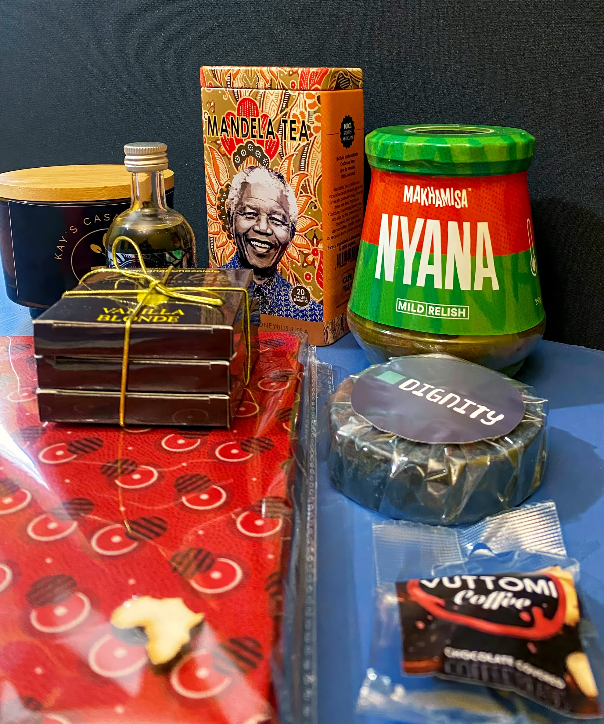 A selection of South African goods on a blue surface, including Mandela Tea, Makhamisa Nyana mild relish, a tin of cashews, olive oil, stacked chocolate bars tied with string, Dignity soap, Vutomi Coffee, and a red patterned cloth.