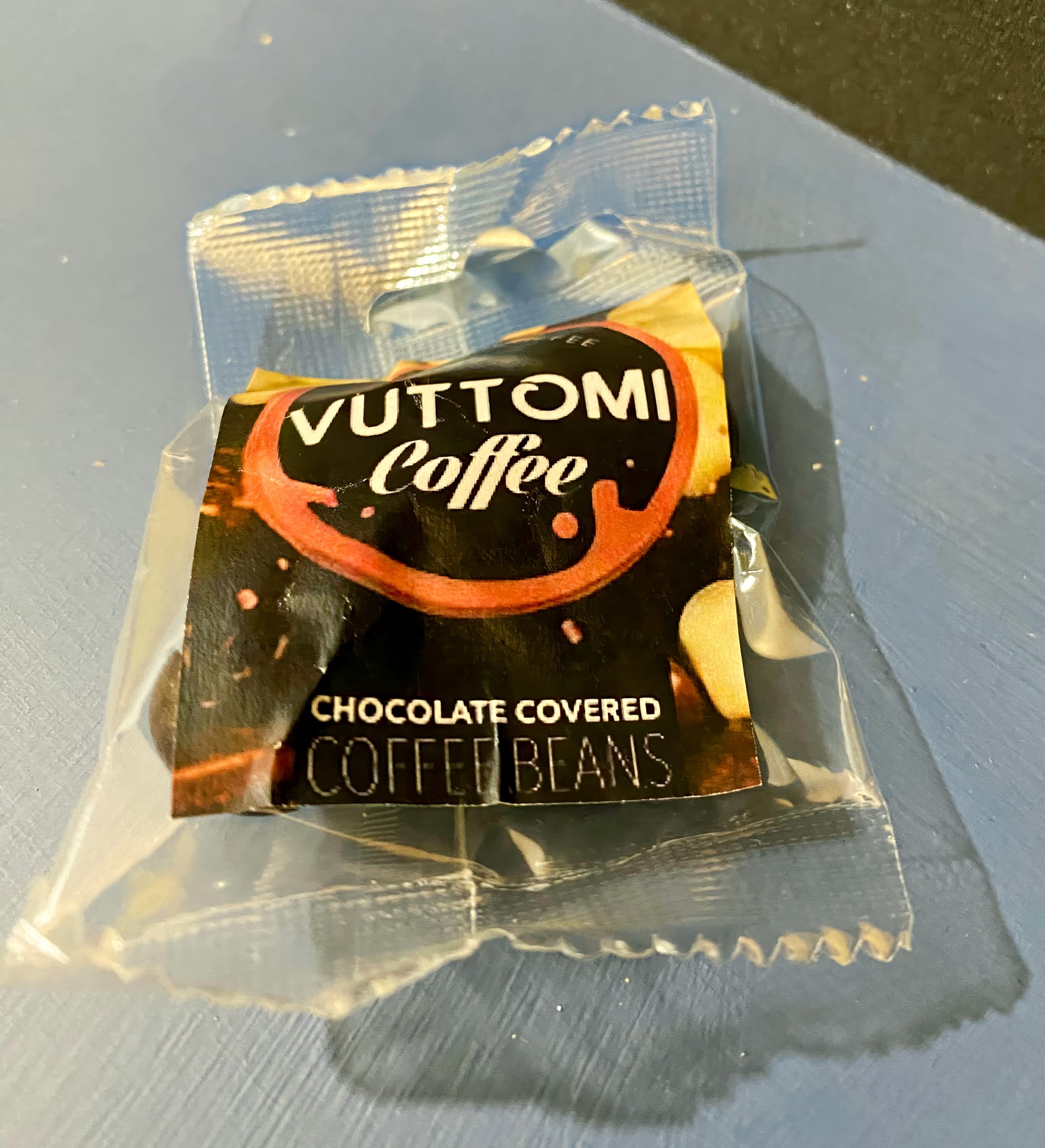 A small clear plastic packet labelled “Vuttomi Coffee” sits on a blue surface. The packaging reads “CHOCOLATE COVERED COFFEE BEANS” with an image of dark chocolate and coffee beans in the background.
