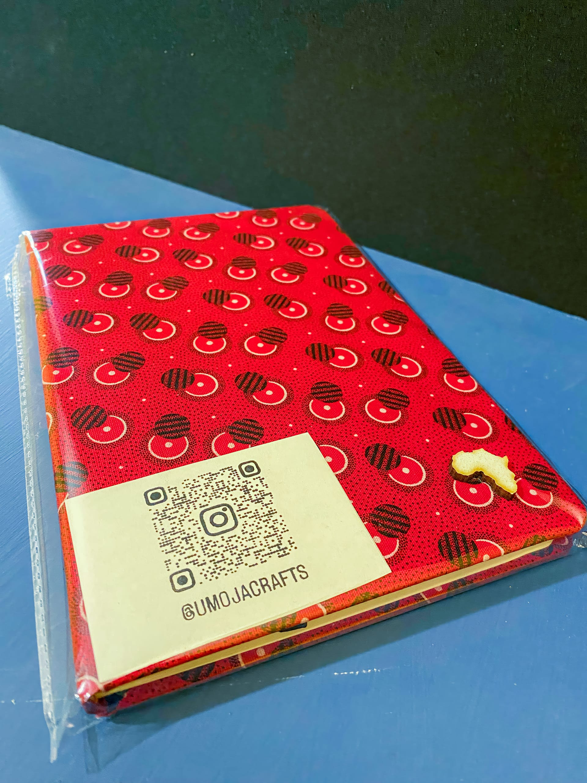 A notebook with a red cover featuring a pattern of black and white shapes lies on a blue surface. A white label with a QR code and the text 