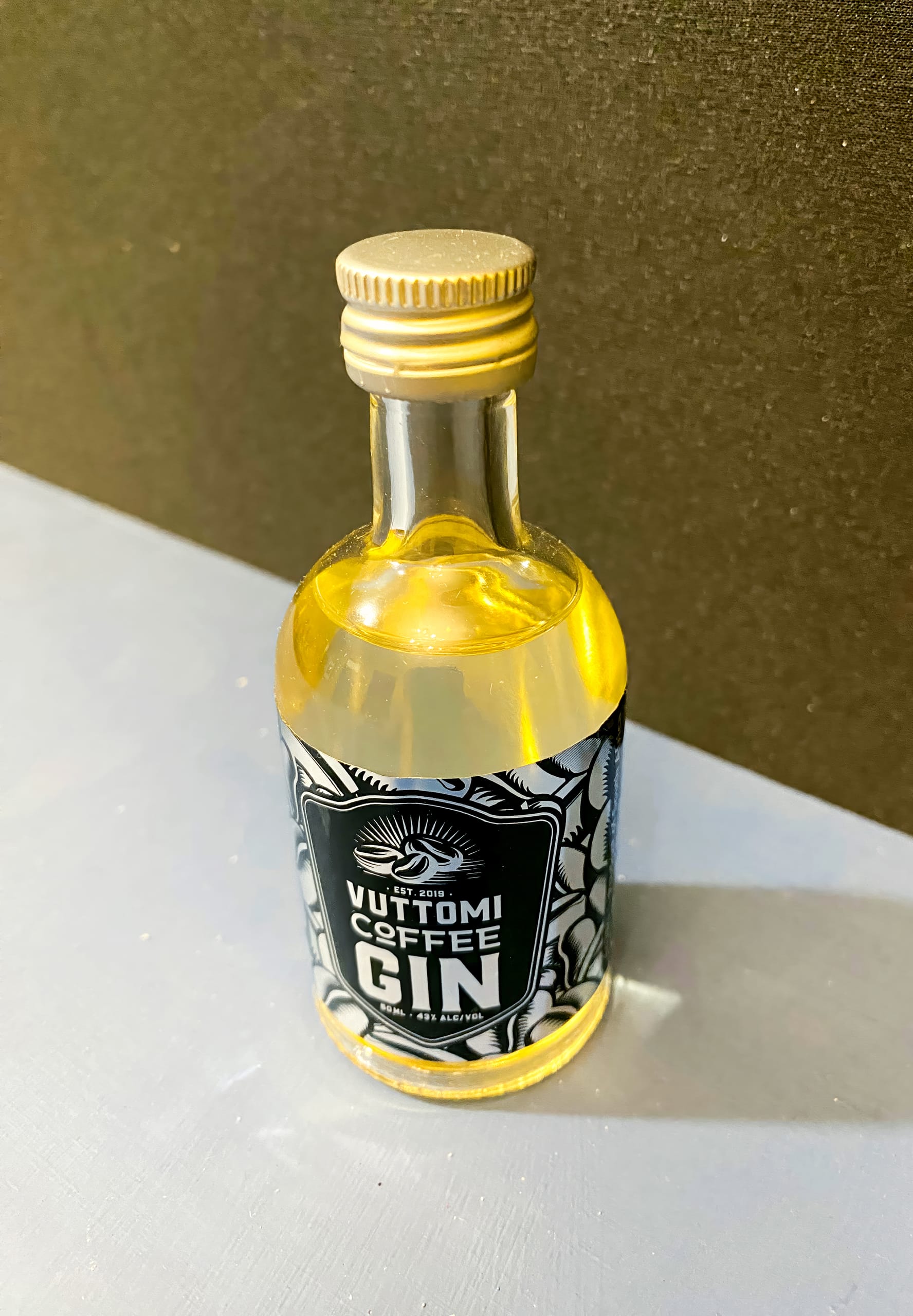 A small clear glass bottle with a golden screw cap contains a yellow liquid. The label reads 