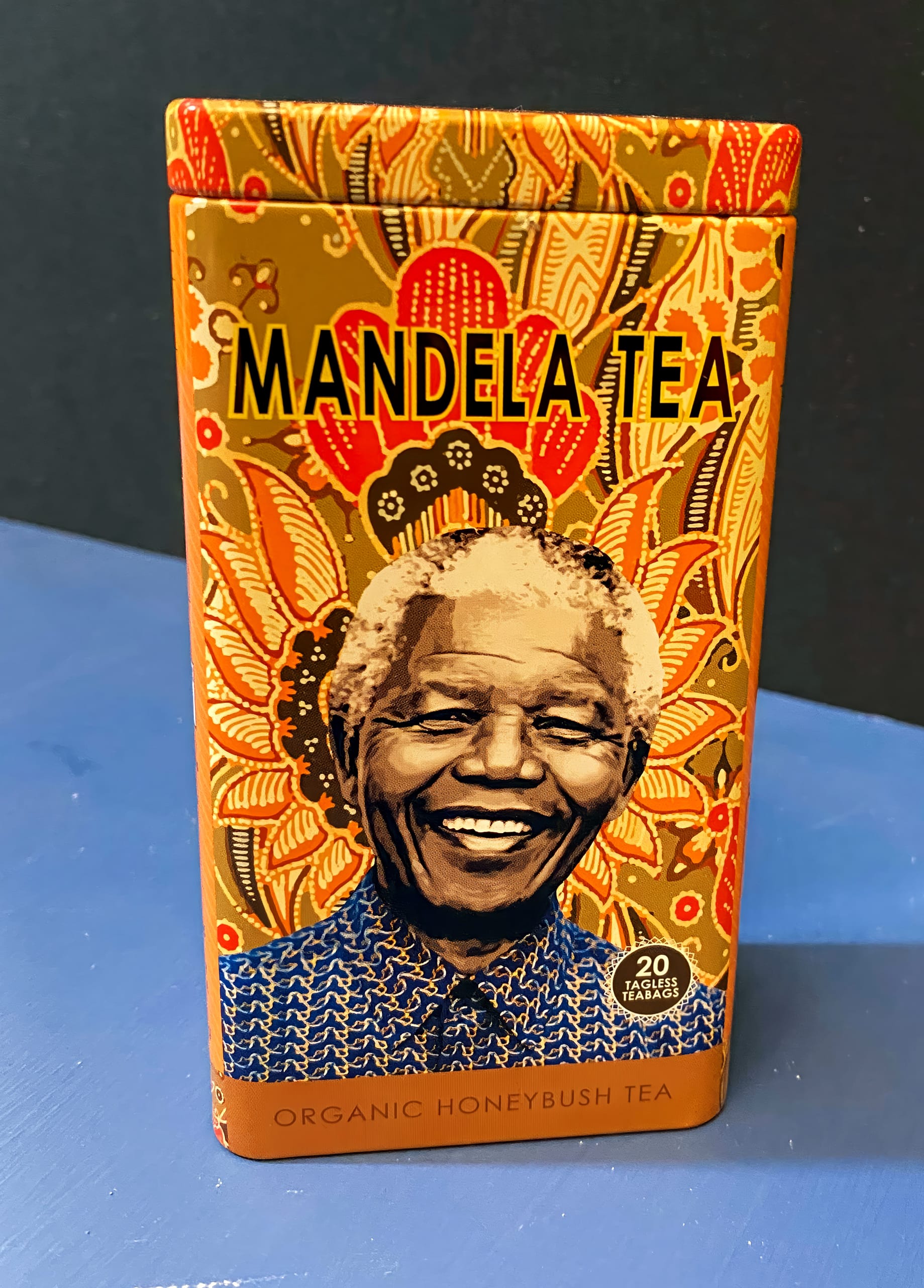 A decorative tin of Mandela Tea features a smiling portrait of Nelson Mandela. The tin is orange with intricate, colourful patterns and text reading 