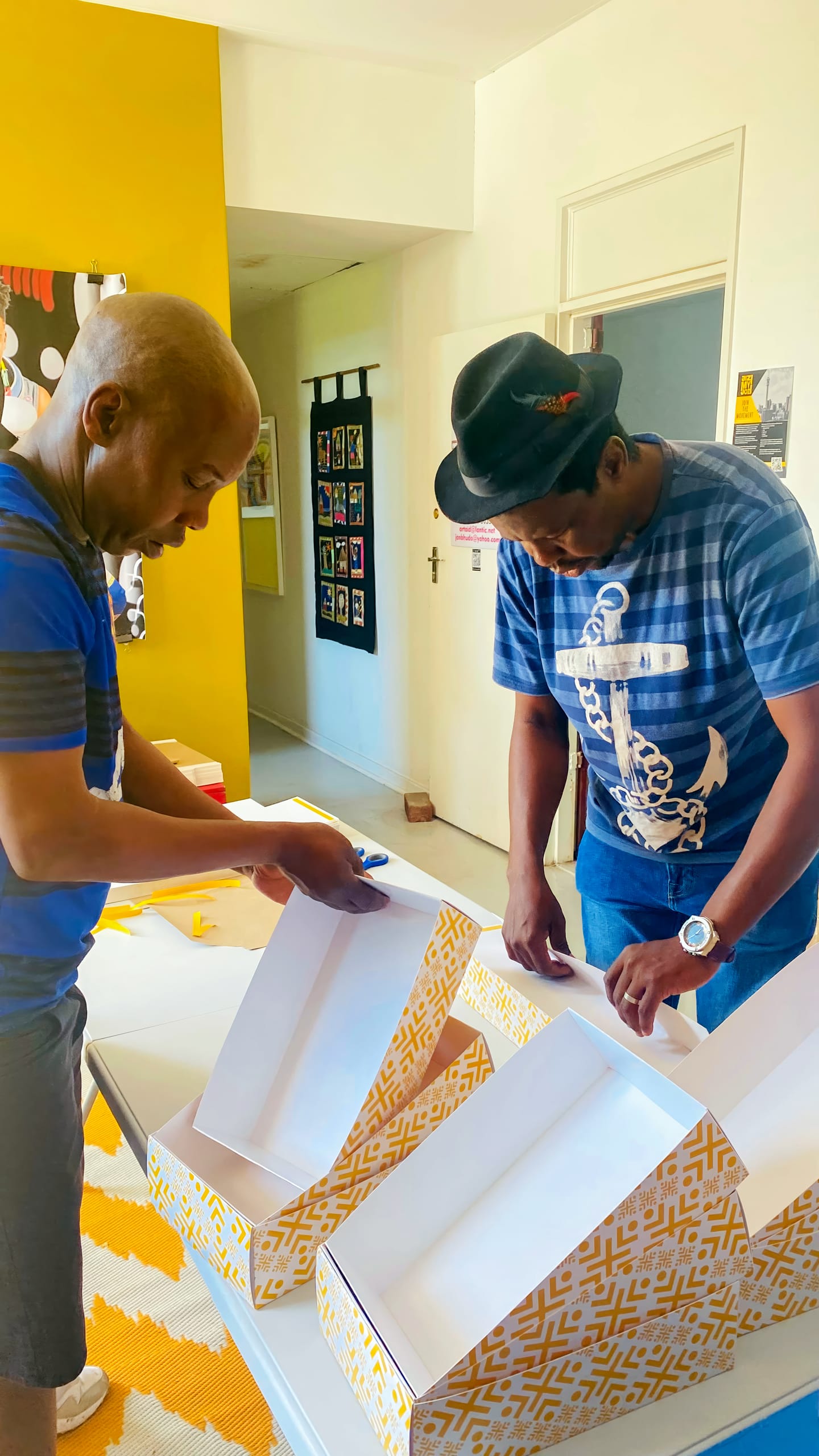 Two men assemble yellow-and-white patterned boxes on a table in a brightly lit room. Both wear blue shirts; one has an anchor design and a hat. Artworks and pictures hang on yellow and white walls in the background. The floor has a patterned rug.