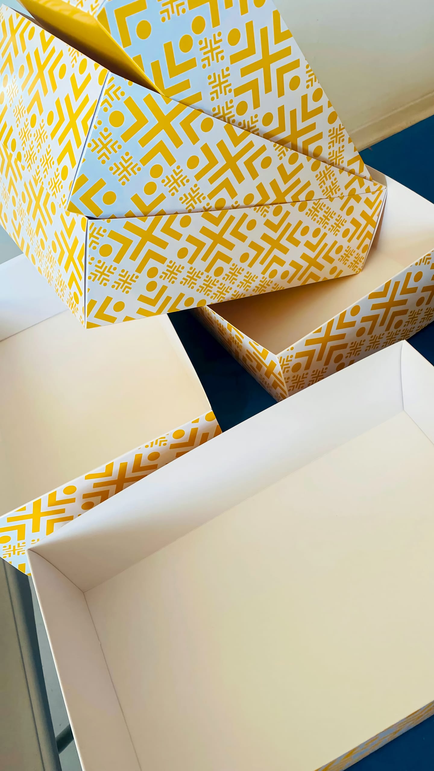 Four empty rectangular boxes with yellow and orange geometric patterns on the outside are arranged on a blue surface. Three boxes are open and nested together, while one is closed and resting on top, showing its patterned lid.