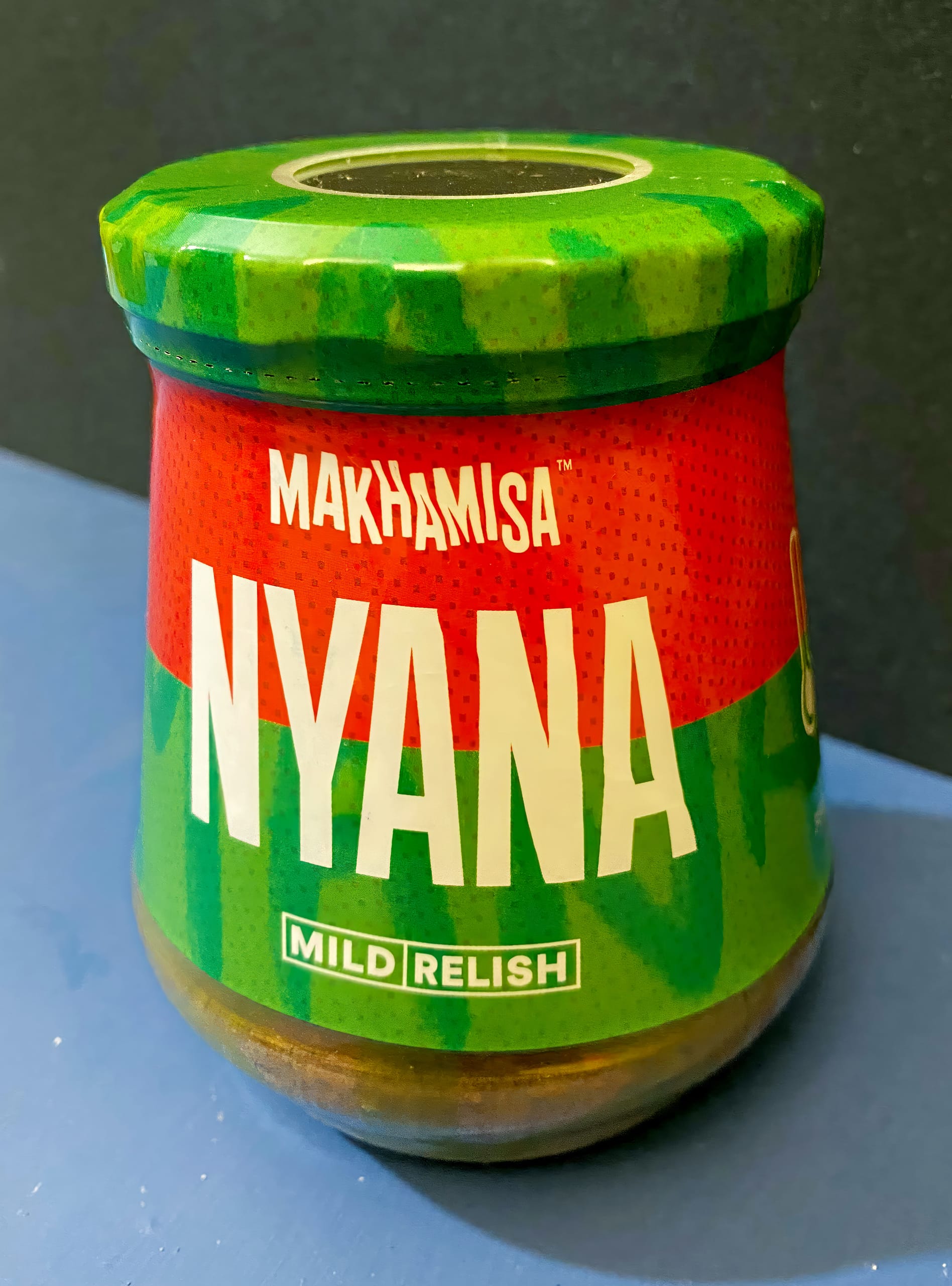 A glass jar with a green lid labelled “MAKHAMISA NYANA MILD RELISH” in large white text. The label is red and green with a bold, modern design. The jar sits on a blue surface against a dark background.