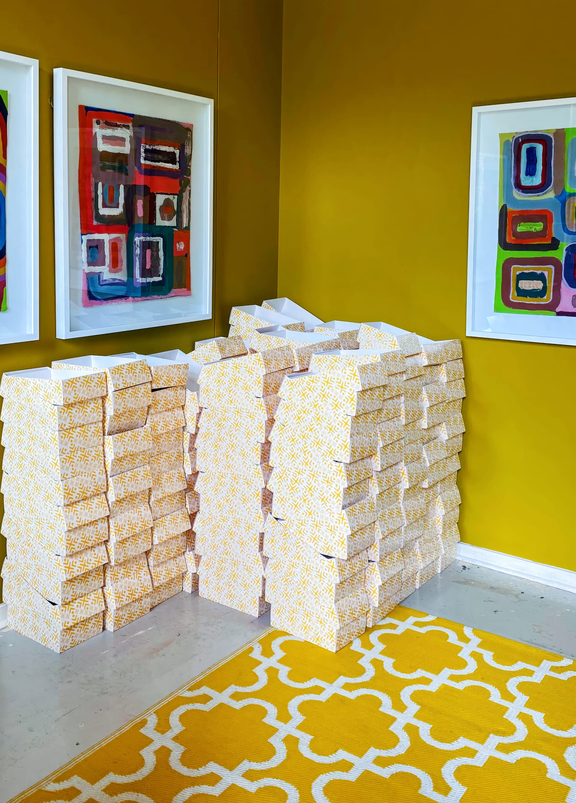 A corner of a vibrant yellow room with patterned yellow and white boxes stacked irregularly. Abstract colourful framed art hangs on the yellow walls, and a matching yellow rug with a white geometric design covers the floor.