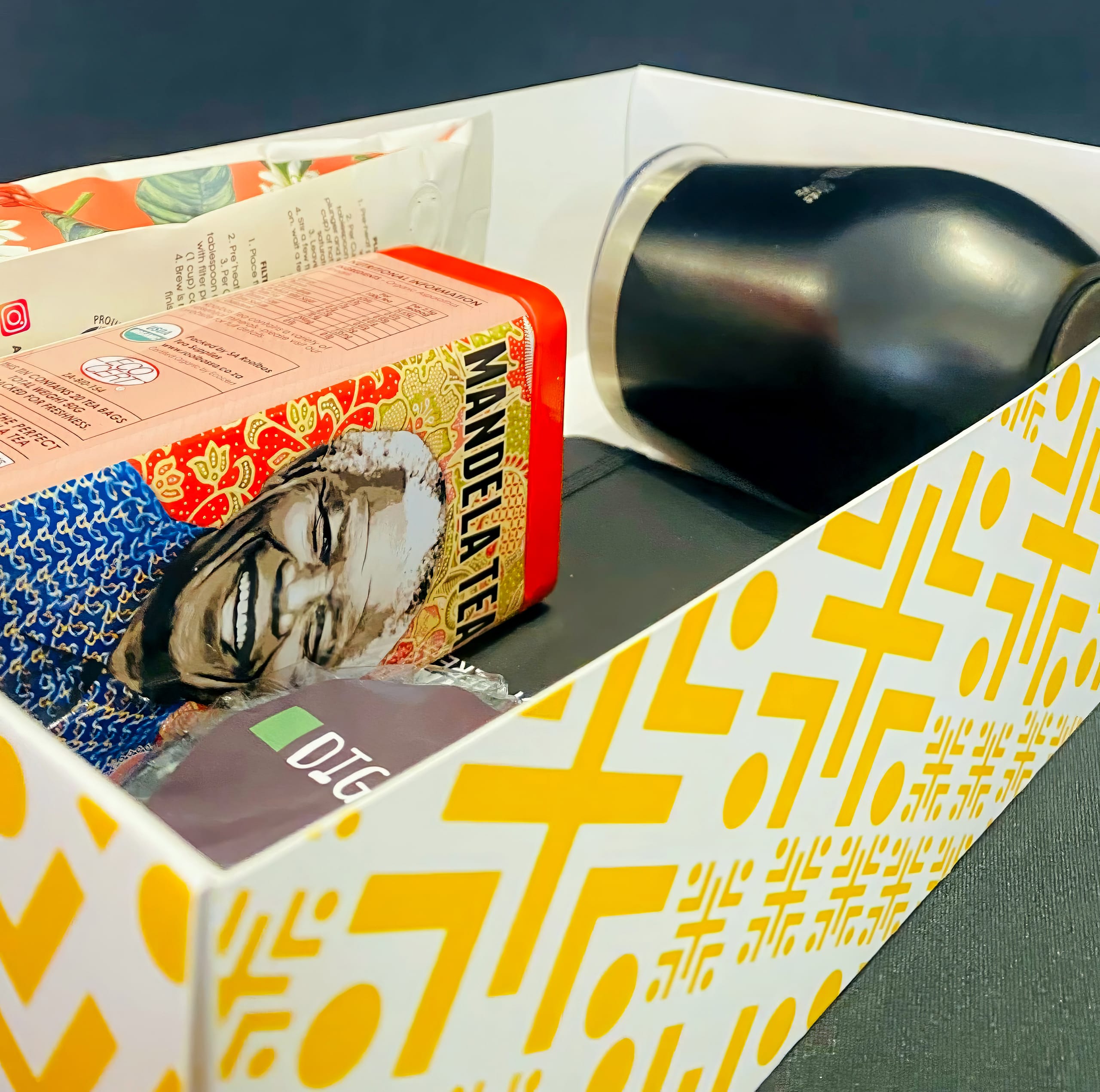A gift box with a yellow geometric pattern on the outside contains a tin of Mandela Tea featuring a smiling portrait, a black tumbler, a packet of OTG coffee, and other partially visible packaged items against a dark background.
