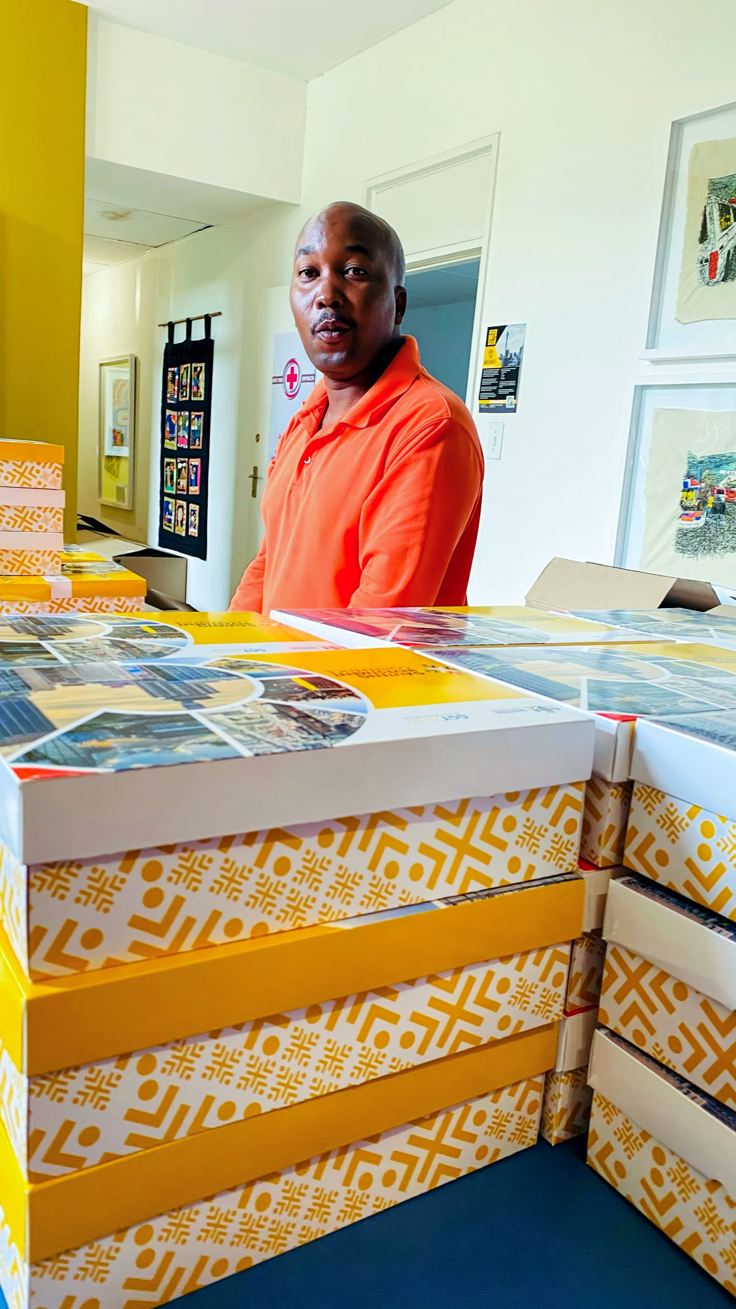 A man in an orange polo shirt stands indoors behind a stack of large, yellow-and-white boxes with geometric patterns. The background features framed artwork on white walls and an open doorway. The man looks slightly towards the camera.