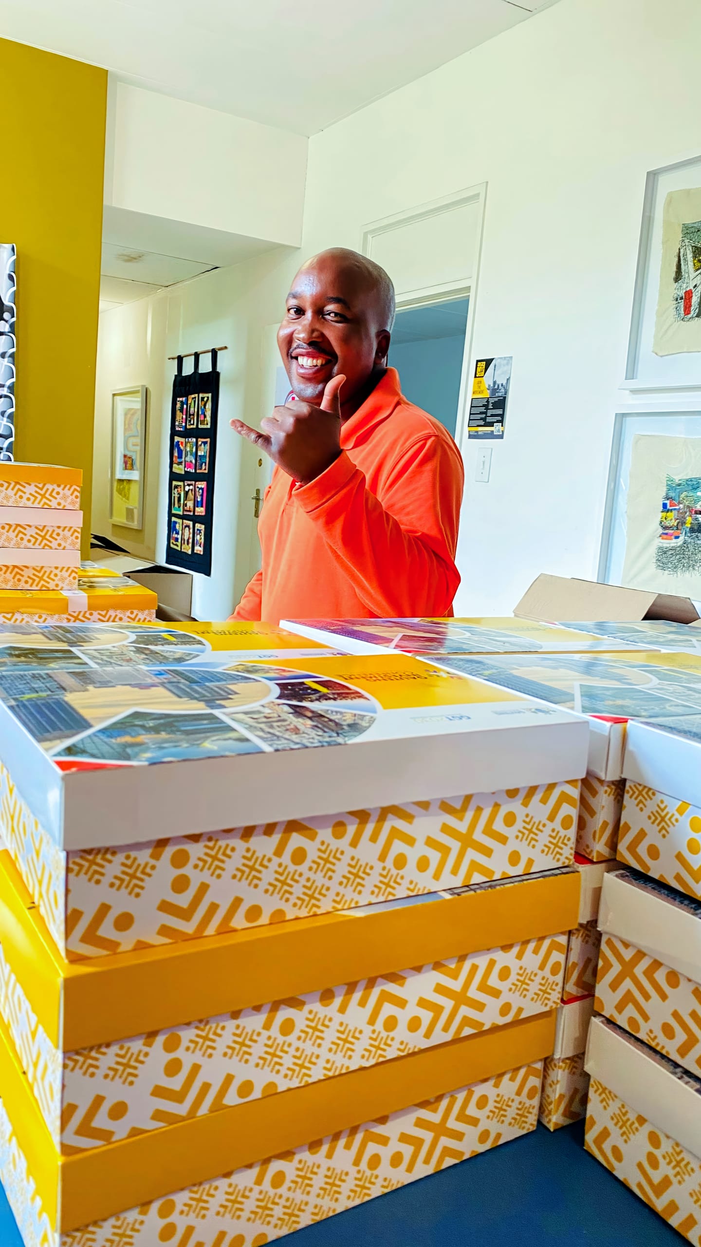 A smiling person in an orange shirt stands behind a table stacked with yellow-and-white patterned boxes. The person holds up a hand in a “hang loose” gesture, with colourful artwork and a yellow accent wall visible in the background.