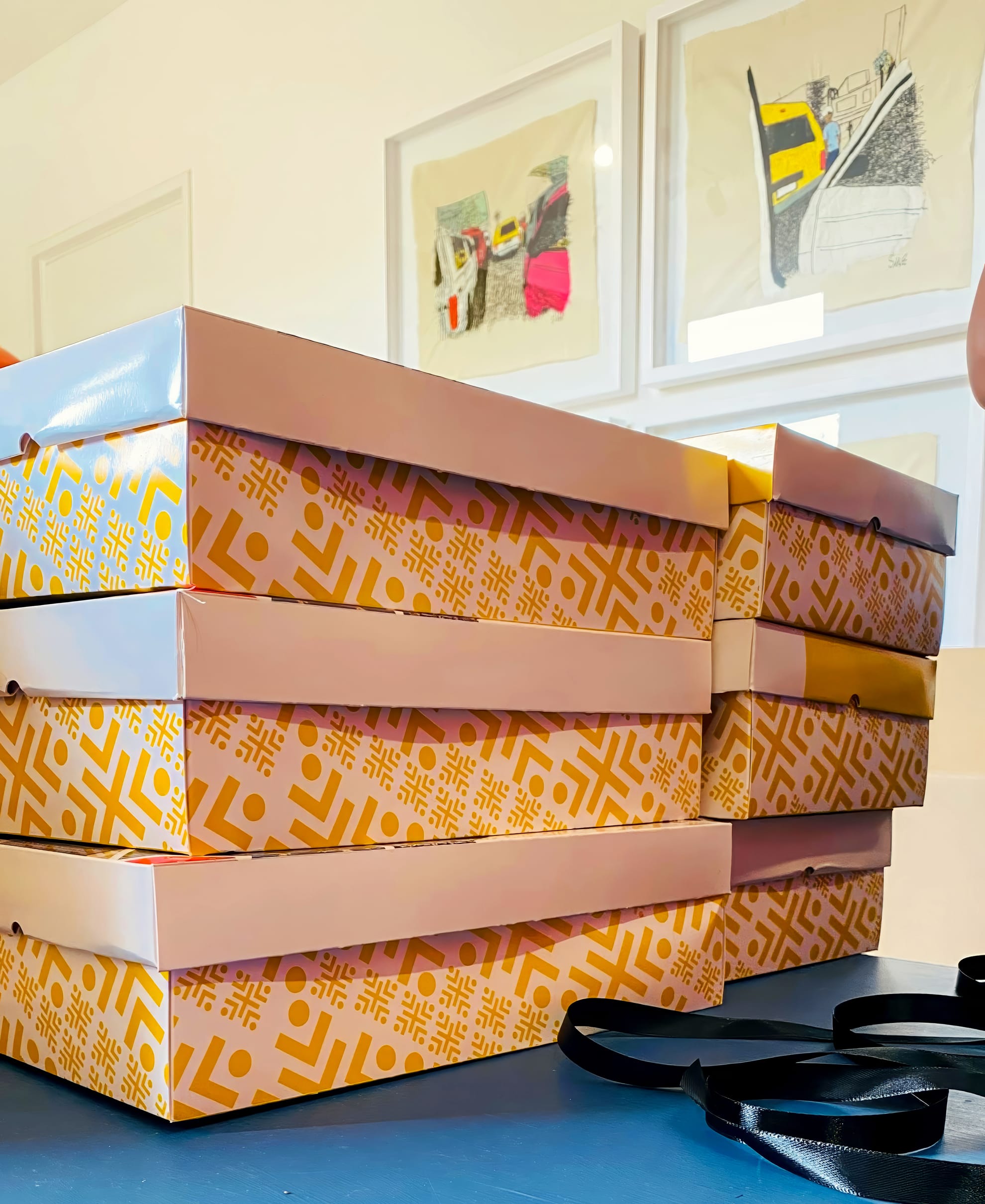 Four yellow boxes with geometric patterns and white lids are stacked on a blue surface. Black ribbon lies in front of the boxes. Framed modern art pieces hang on the white wall in the background. The scene is brightly lit.
