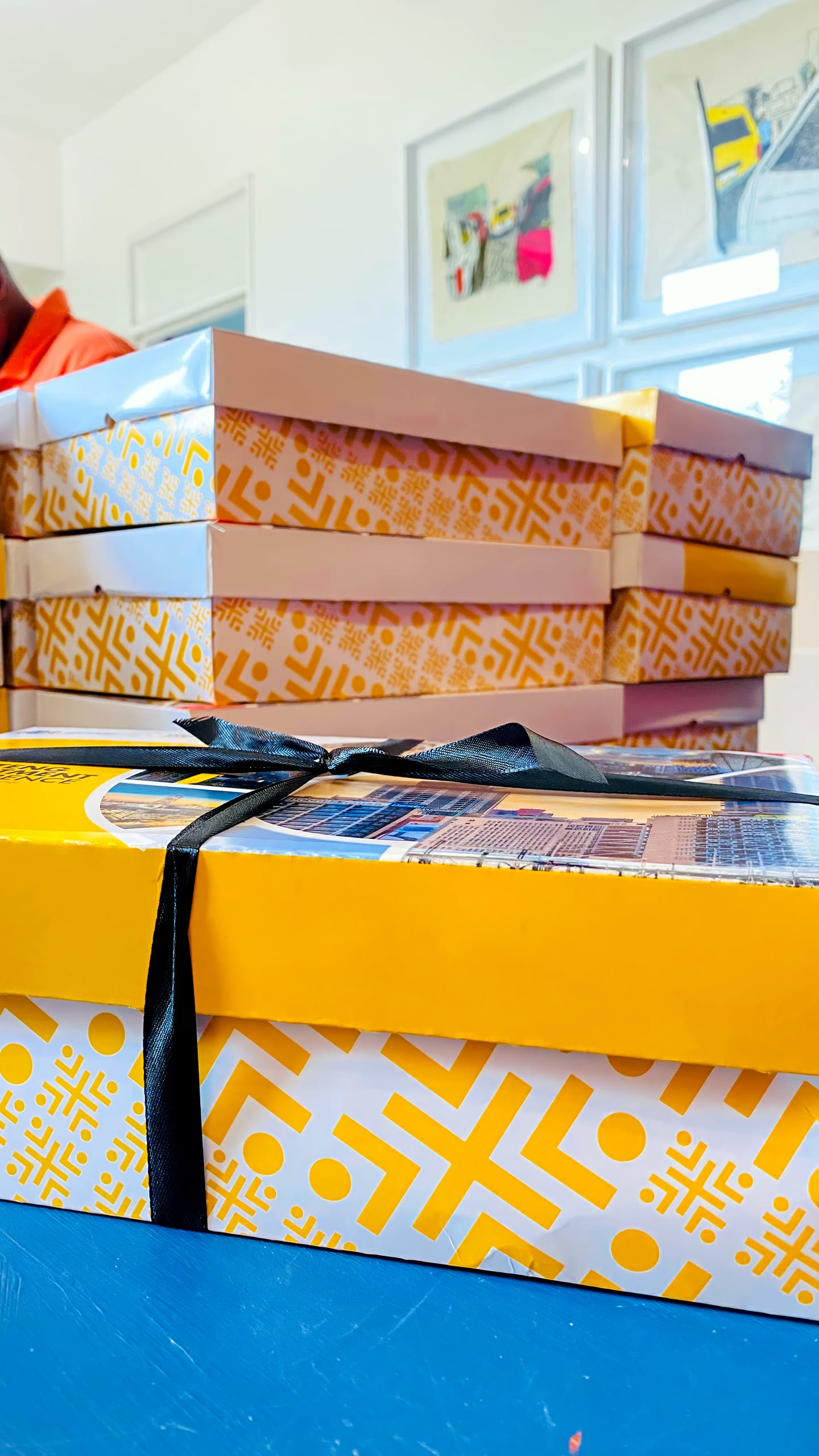 A stack of decorative yellow and white patterned boxes rests on a table, with one box in the foreground tied with a black ribbon. The background shows framed artwork on a white wall and part of a person out of focus.