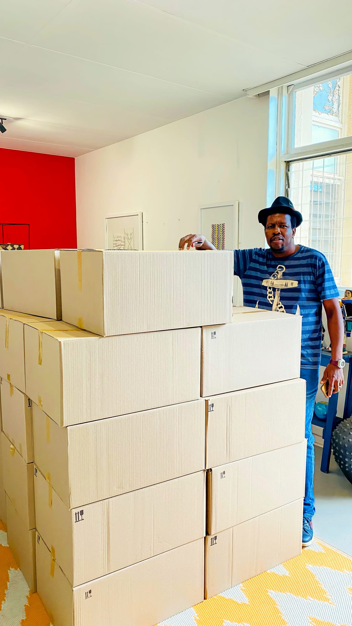 A man in a striped shirt and hat stands indoors behind a large stack of sealed cardboard boxes. The boxes are arranged in three rows. The room has a red feature wall, artworks on white walls, and sunlight comes through a tall window.