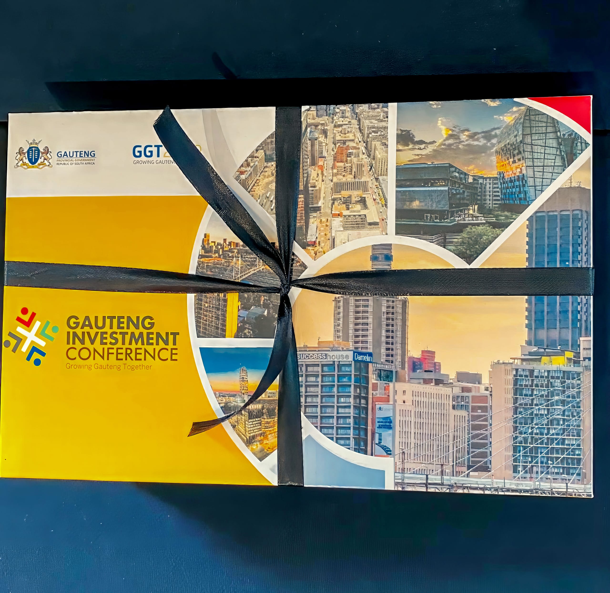 A rectangular gift box tied with a black ribbon features cityscape photos and the logo for the Gauteng Investment Conference. The bottom left area is yellow with the conference name and slogan “Growing Gauteng Together.” Skyscrapers and buildings are visible in the images.