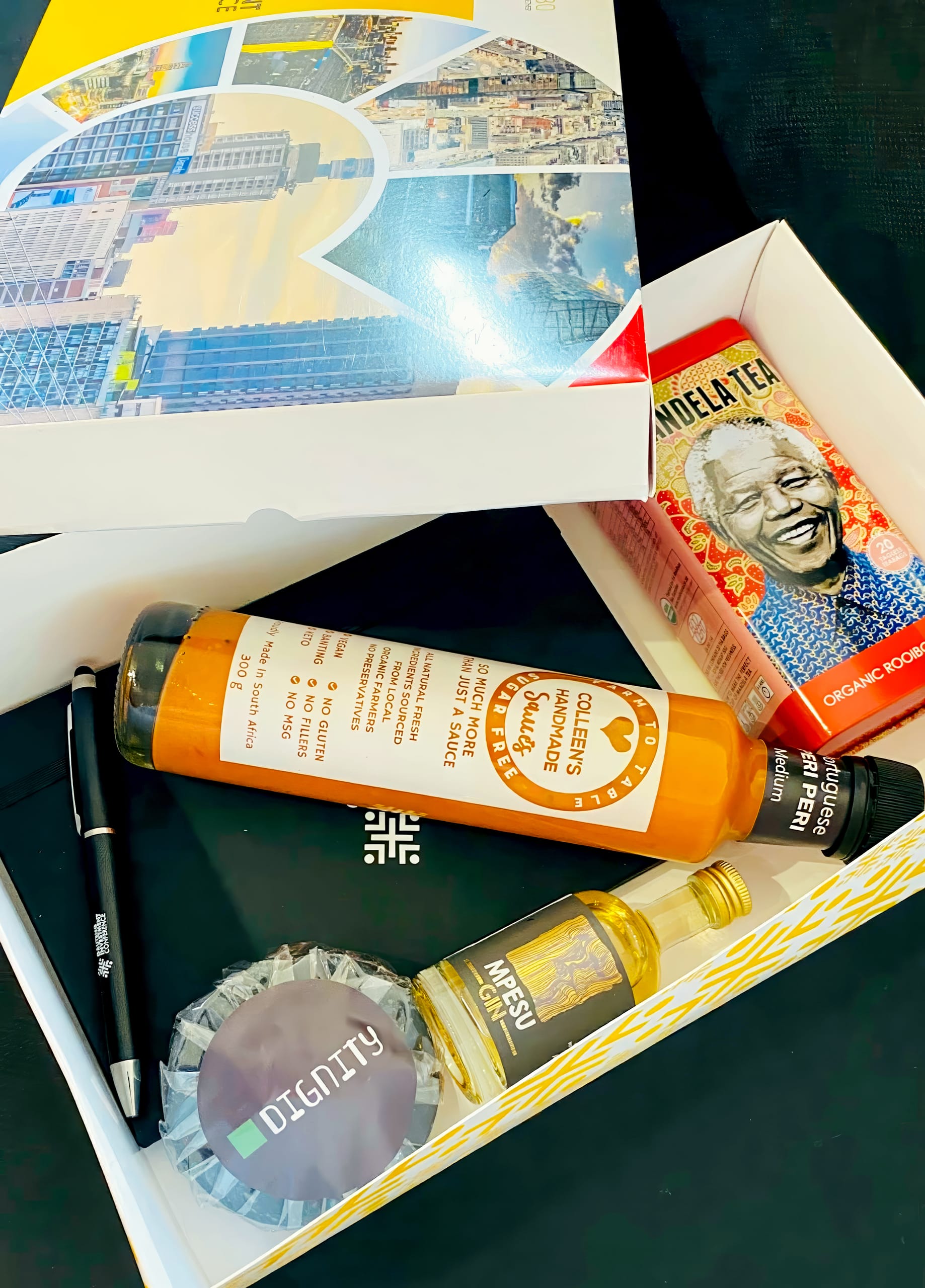 A gift box contains Mandela Tea, a Collerette Moutarde sauce bottle, a mini Marsu Cognac bottle, a black notebook with a white geometric logo, a round black item labelled 