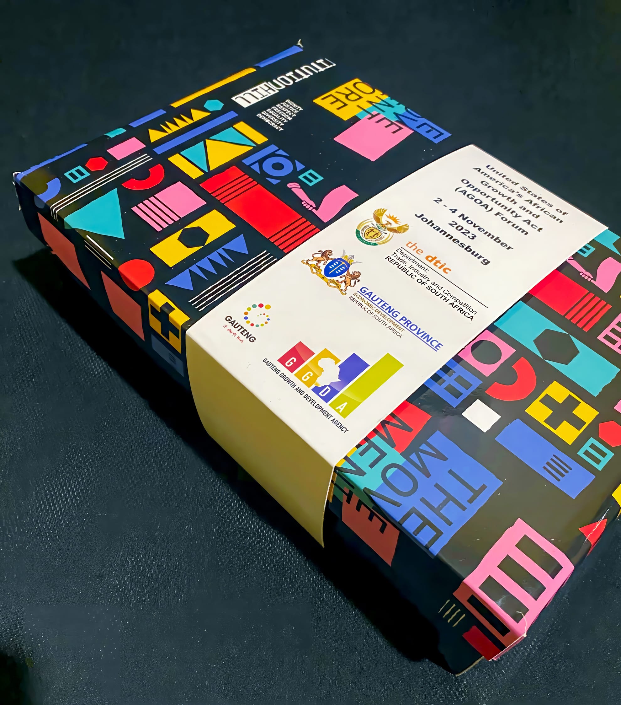 A rectangular box with a colourful geometric pattern and a white band displaying multiple logos, including South African government and Gauteng Province identities, lies on a dark surface. The box looks official and vibrant.