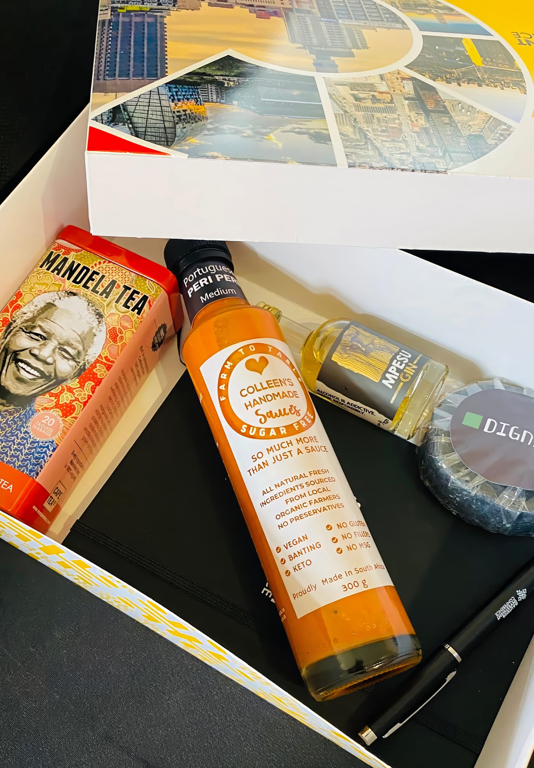A gift box contains Mandela Tea, Colleen’s Handmade Peri Peri Sauce, a small bottle of liquid, a round DGNN-branded soap, a black face mask, and a black pen on a white surface with colourful cityscape images on the box lid.