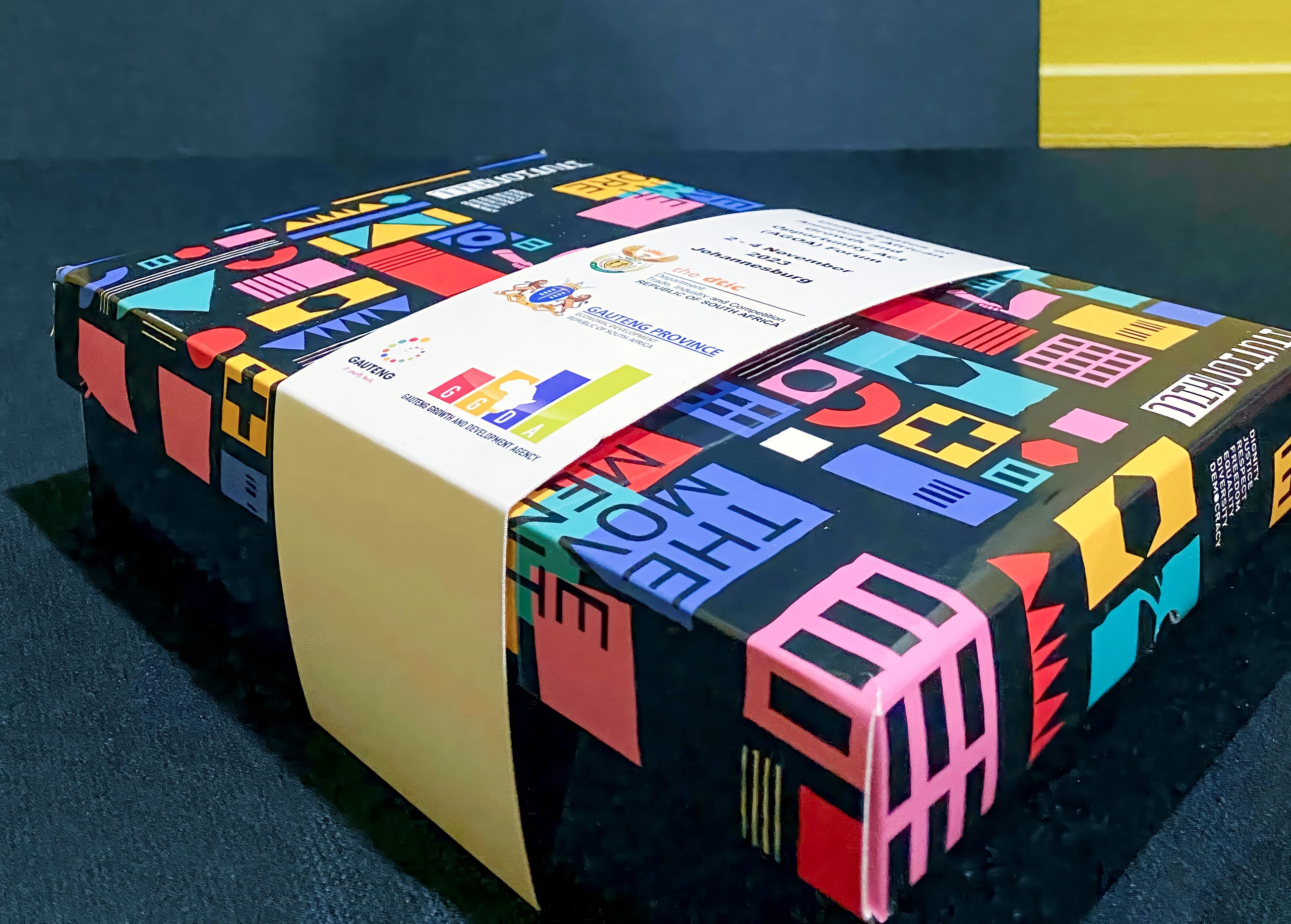 A rectangular box wrapped in colourful, geometric-patterned paper, featuring shapes and symbols in red, blue, yellow, and pink. A white band with logos and text in Thai is wrapped around the box, which sits on a dark textured surface.