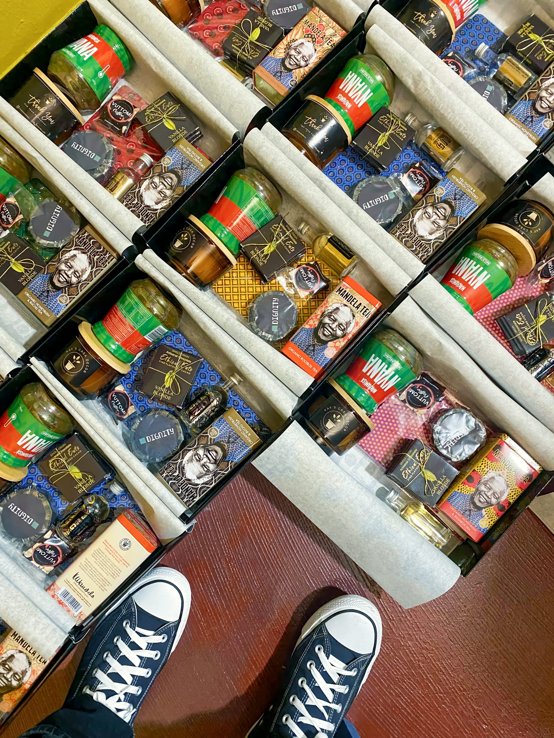 Several open gift boxes filled with various luxury grooming products, colourful ties, and jars, are arranged neatly on a reddish-brown wooden floor. The tip of a person's black-and-white trainers is visible at the bottom of the frame.
