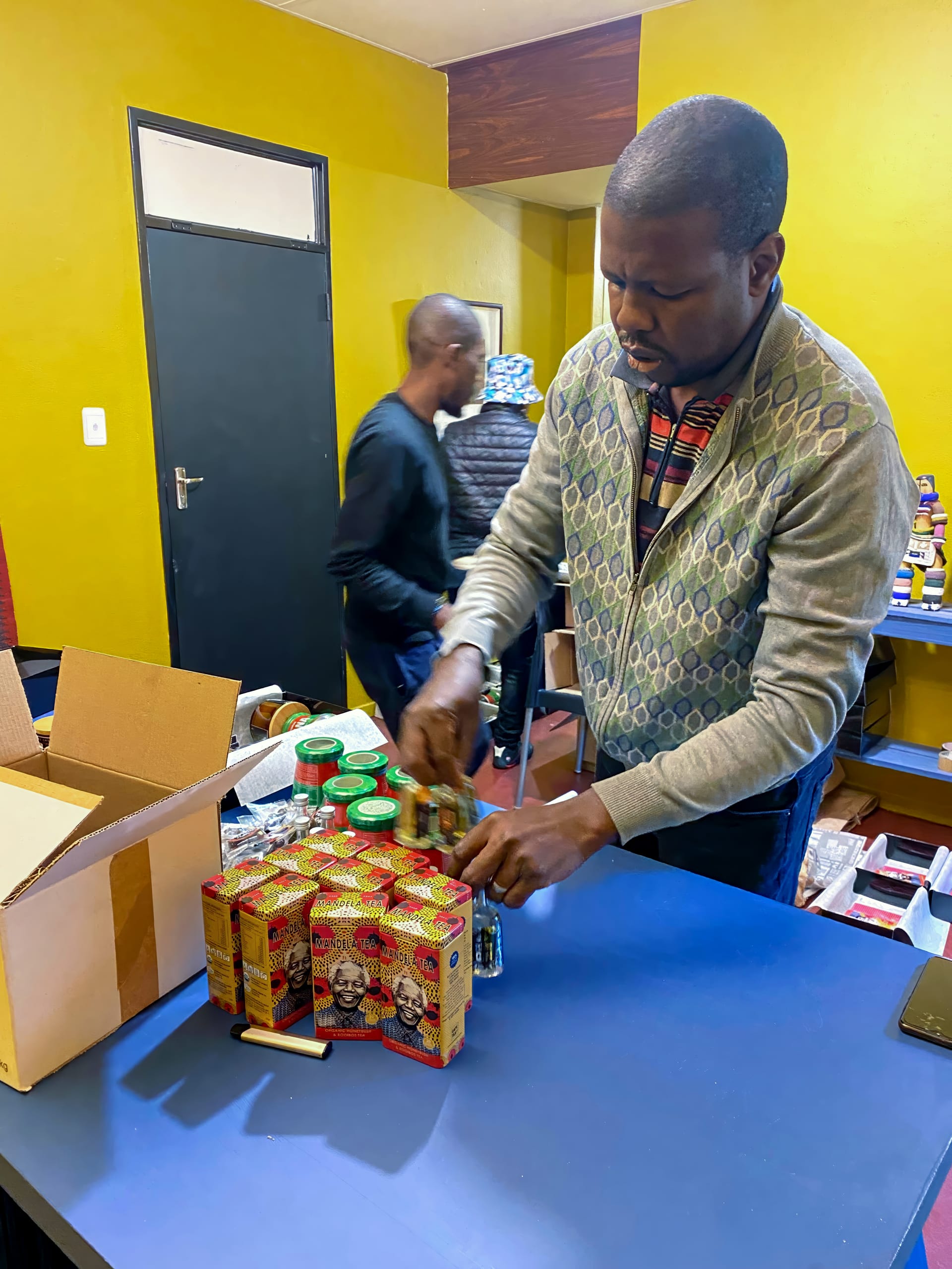 A man in a grey jumper with patterned sleeves arranges jars and stacked boxes of food on a blue table. An open cardboard box sits nearby. Another person in a black shirt is blurred in the background, walking past yellow walls and shelves.