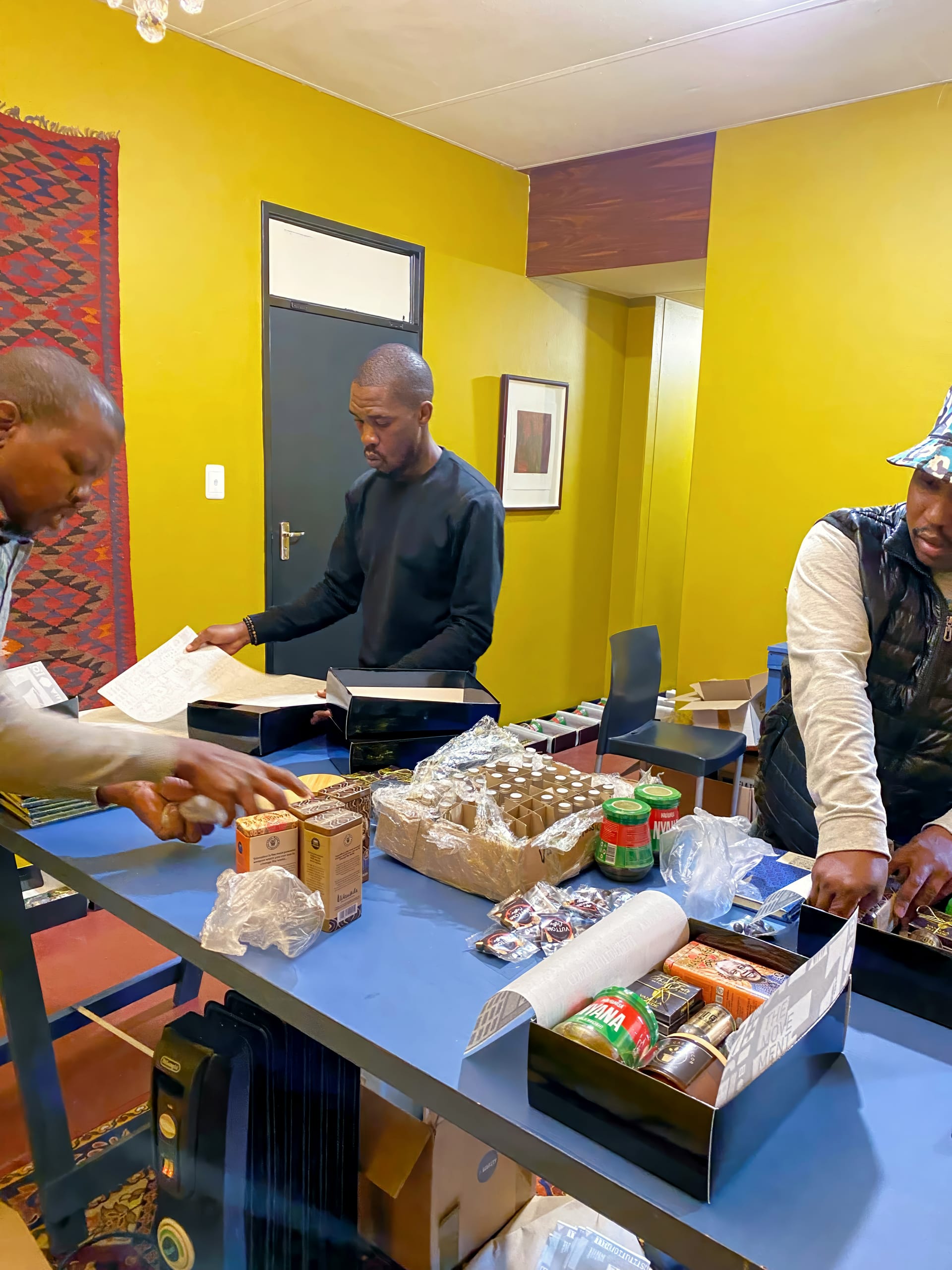 Three men stand at a blue table in a yellow-walled room, organising various food items, tins, jars, and papers. Shelves hold more supplies. The scene suggests teamwork and preparation, possibly for food distribution or community aid.