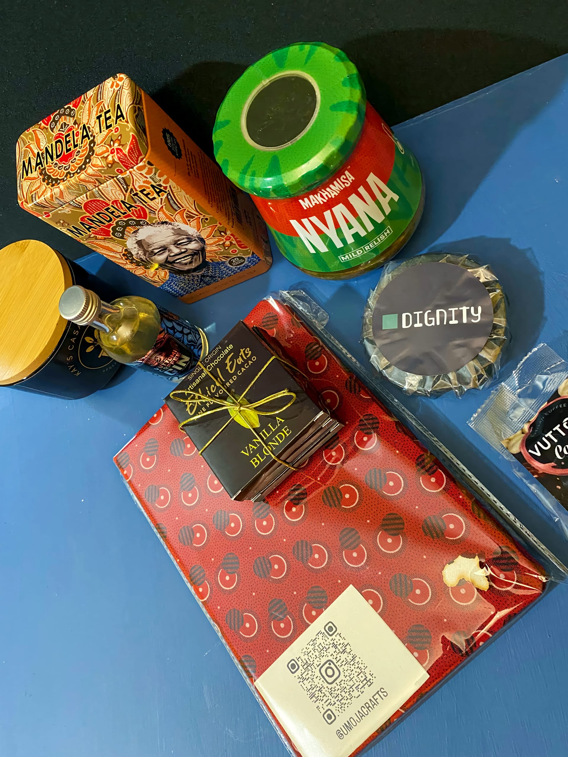 A variety of products on a blue surface: Mandela Tea tin, a green-lidded Nyana container, a small bottle of oil, a black and gold vanilla candle, a red cloth with a QR code, a soap labelled “DIGNITY,” and a wrapped chocolate labelled “NUTRITION.”.