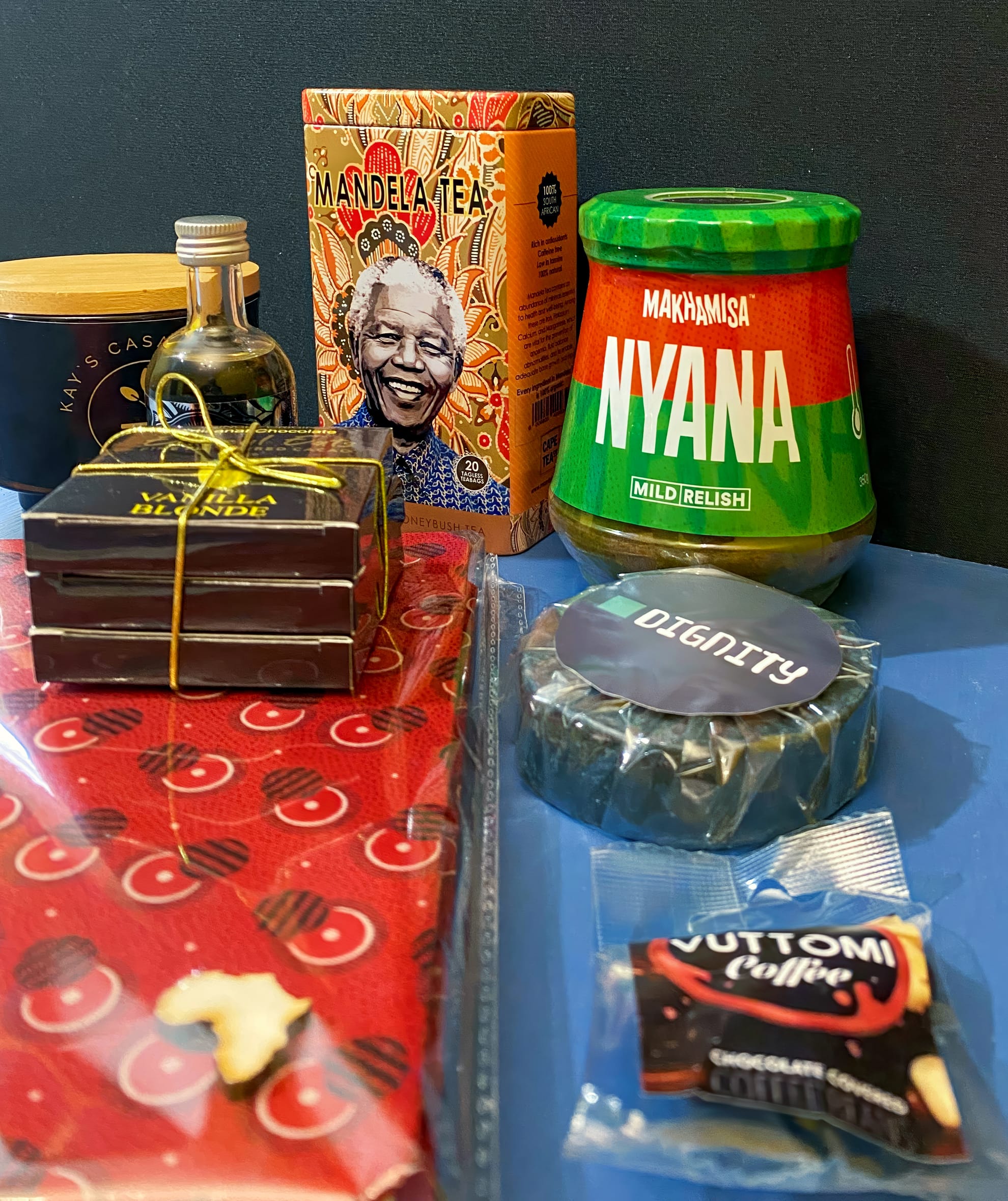 A colourful assortment of South African products: Mandela Tea box, Makhamisa Nyana mild relish jar, a bottle of olive oil, stacked boxes of chocolates, a packet of Bottom Coffee, a wrapped cheese wheel, and a red patterned cloth or tie.