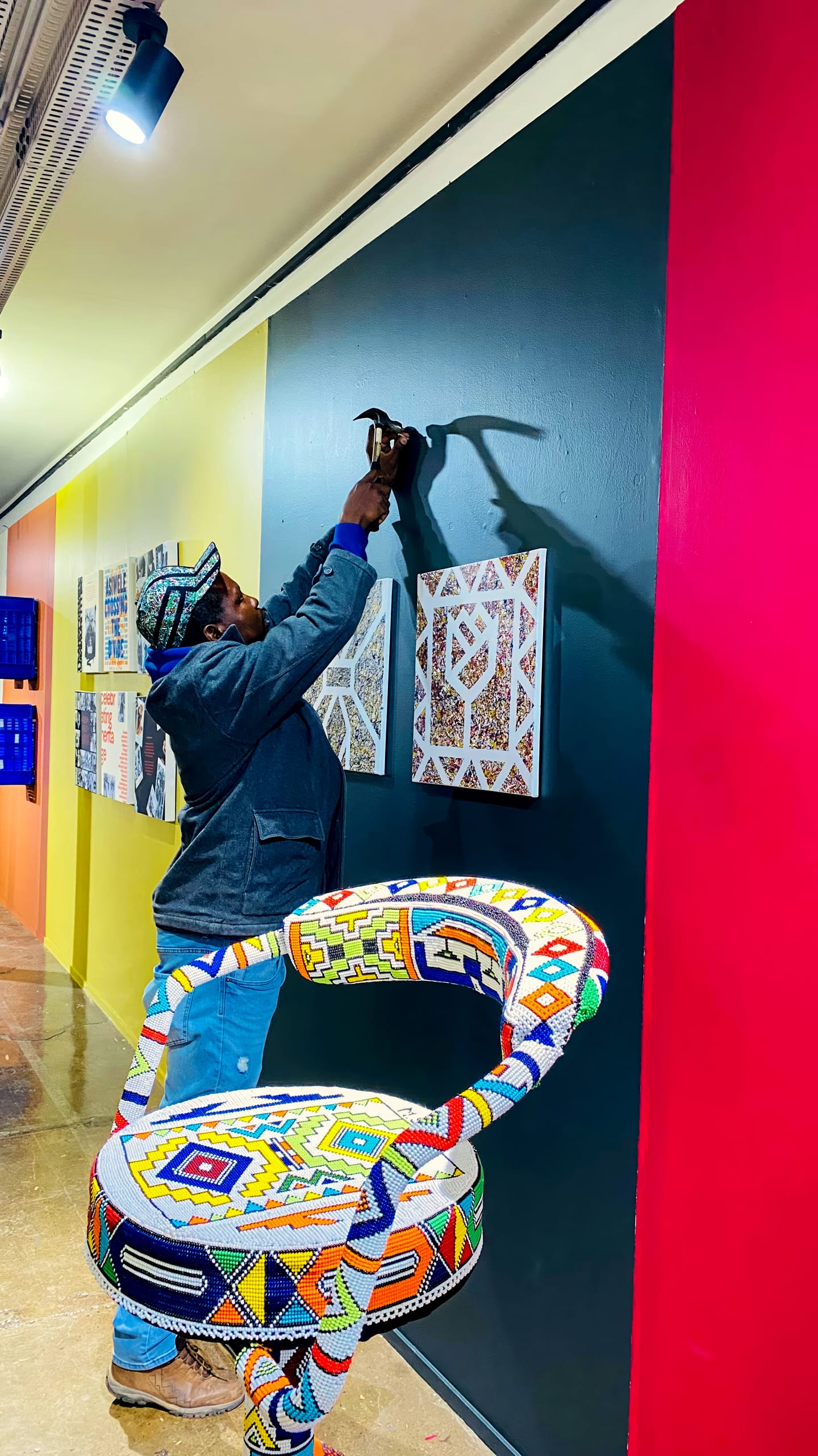 A person in a colourful hat and jacket hangs artwork on a dark teal gallery wall using a hammer. The wall features geometric-patterned art, and in the foreground is a vibrant, beaded chair with intricate, multicoloured patterns. The floor is shiny and tan.