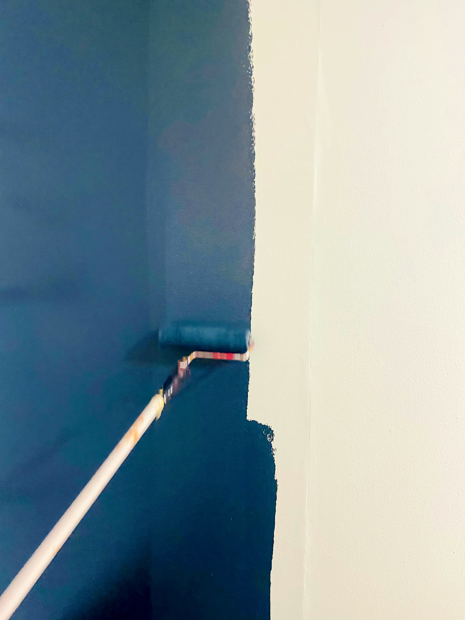 A paint roller on a pole is being used to apply dark blue paint to a light-coloured wall. The roller is halfway up the wall, creating a clear dividing line between the freshly painted dark blue area and the unpainted light area.