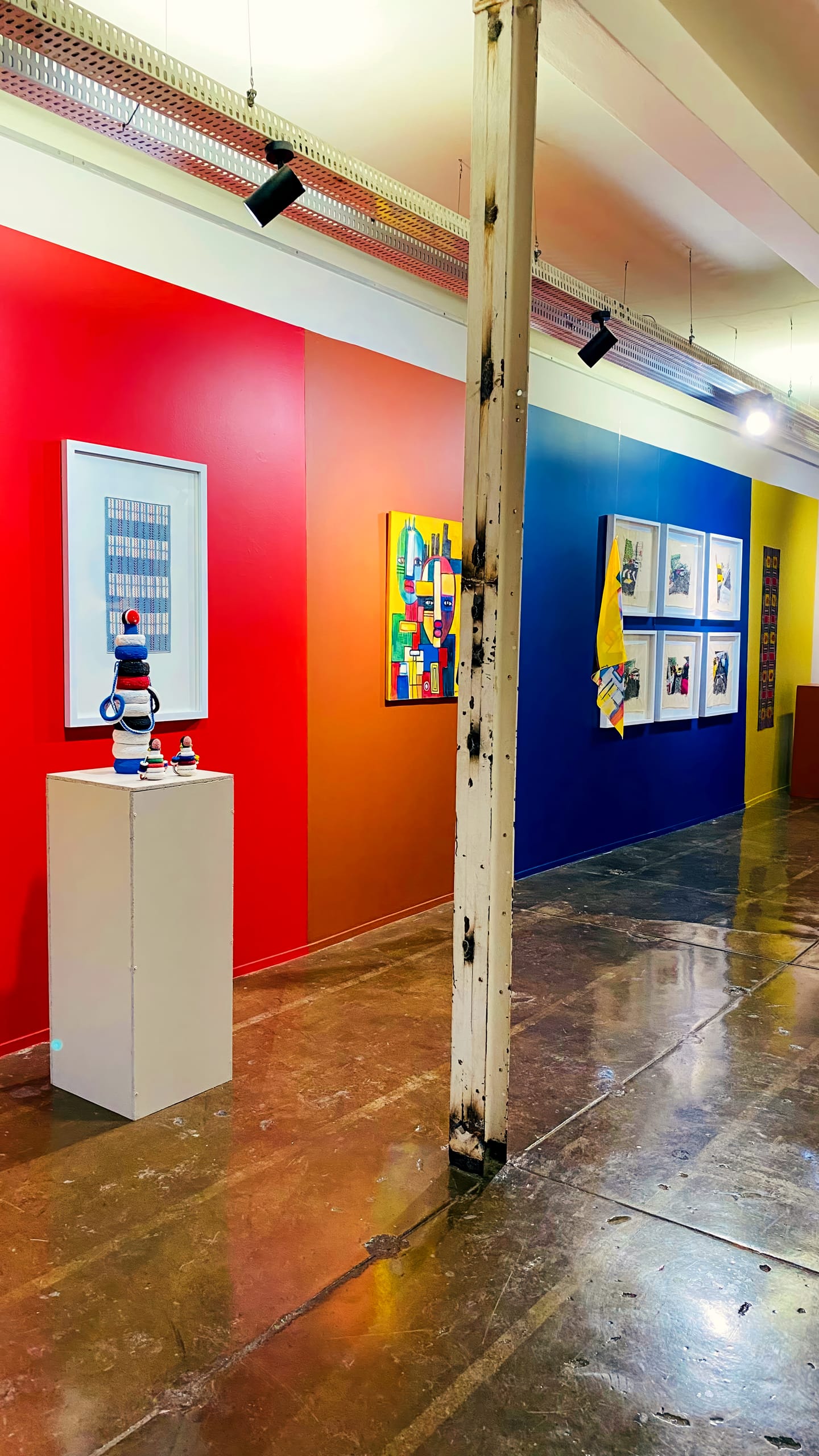 A vibrant art gallery features a red wall with framed artwork and a sculpture on a white plinth, transitioning into an orange and blue wall displaying colourful paintings and photographs. A worn white column stands in the centre on the polished floor.