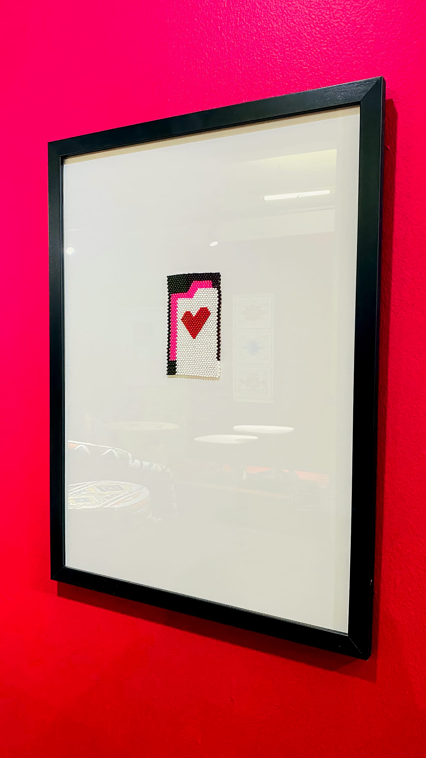A framed artwork hangs on a bright pink wall. The artwork features a minimalist pixelated design of a folder icon with a red heart in the centre, set against a white background and surrounded by a black frame.