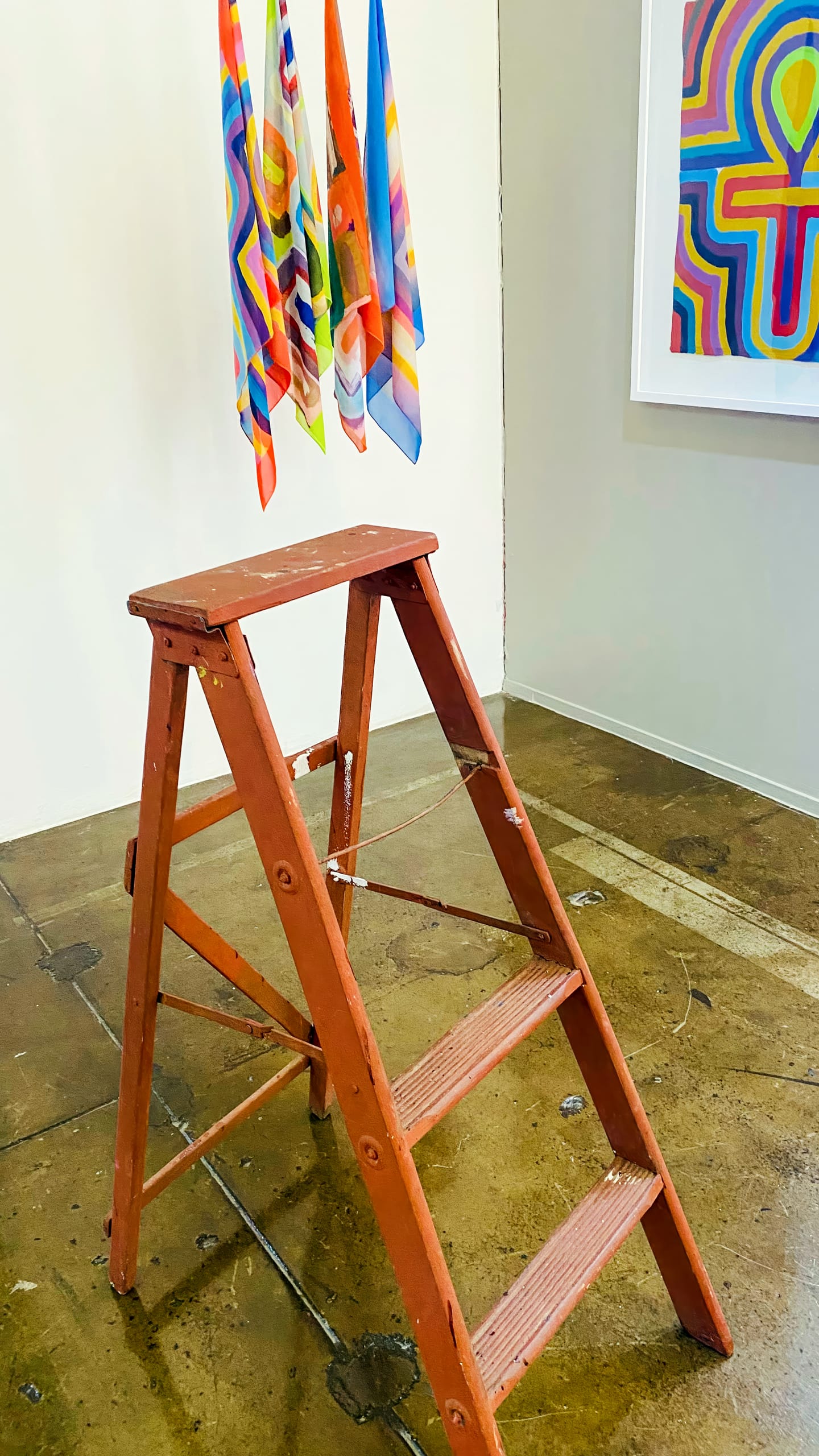 A small, red, wooden stepladder stands on a worn, brown concrete floor. Behind it hang several colourful, geometric-patterned cloths, with a framed abstract artwork of similar patterns on the white wall to the right.