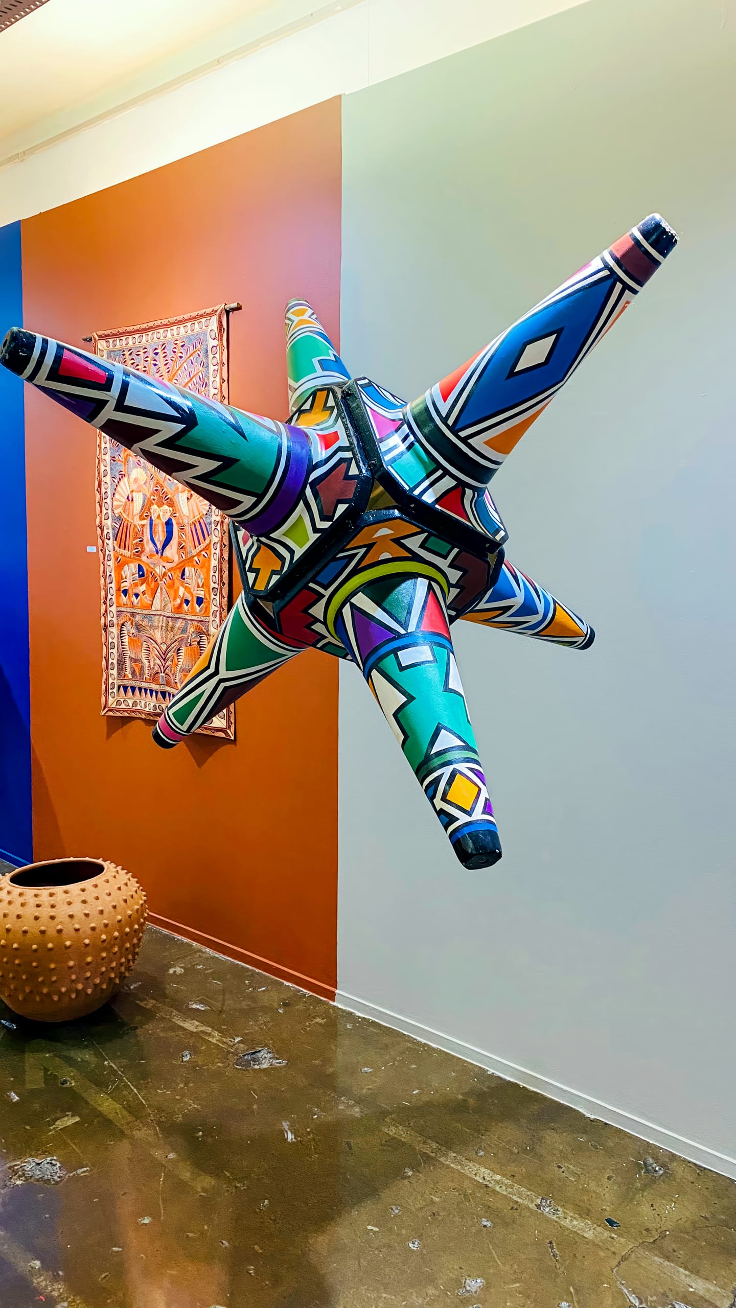 A large, multicoloured geometric star sculpture with six points is suspended in a gallery. Behind it, a patterned textile hangs on an orange and white wall. A textured, round tan vessel sits on the concrete floor to the left. The scene is vibrant and modern.