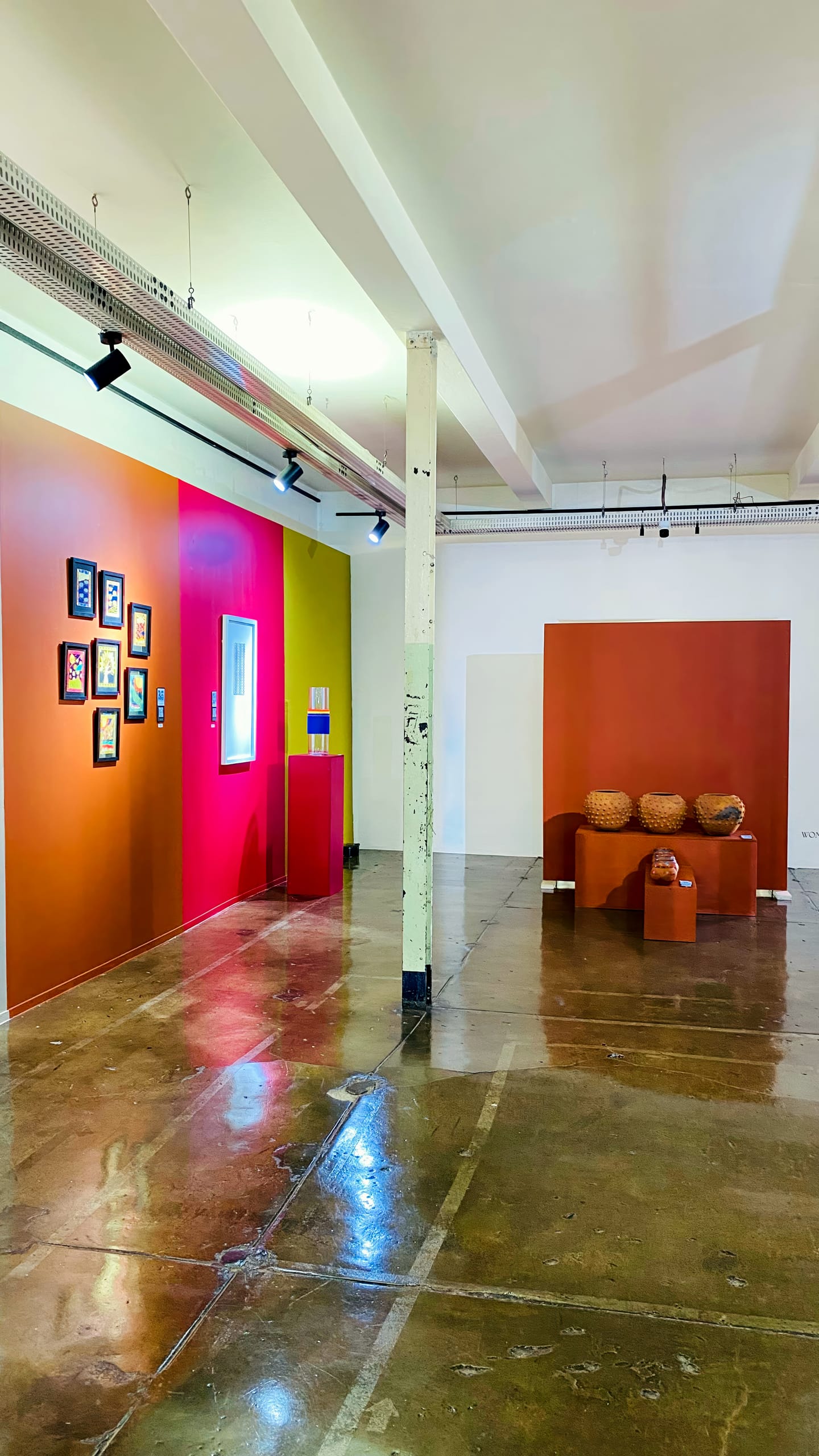 A modern art gallery with shiny concrete floors, colourful walls in orange and pink tones, small framed artworks on the left, and woven baskets displayed on plinths to the right. A white pillar stands in the centre of the space.