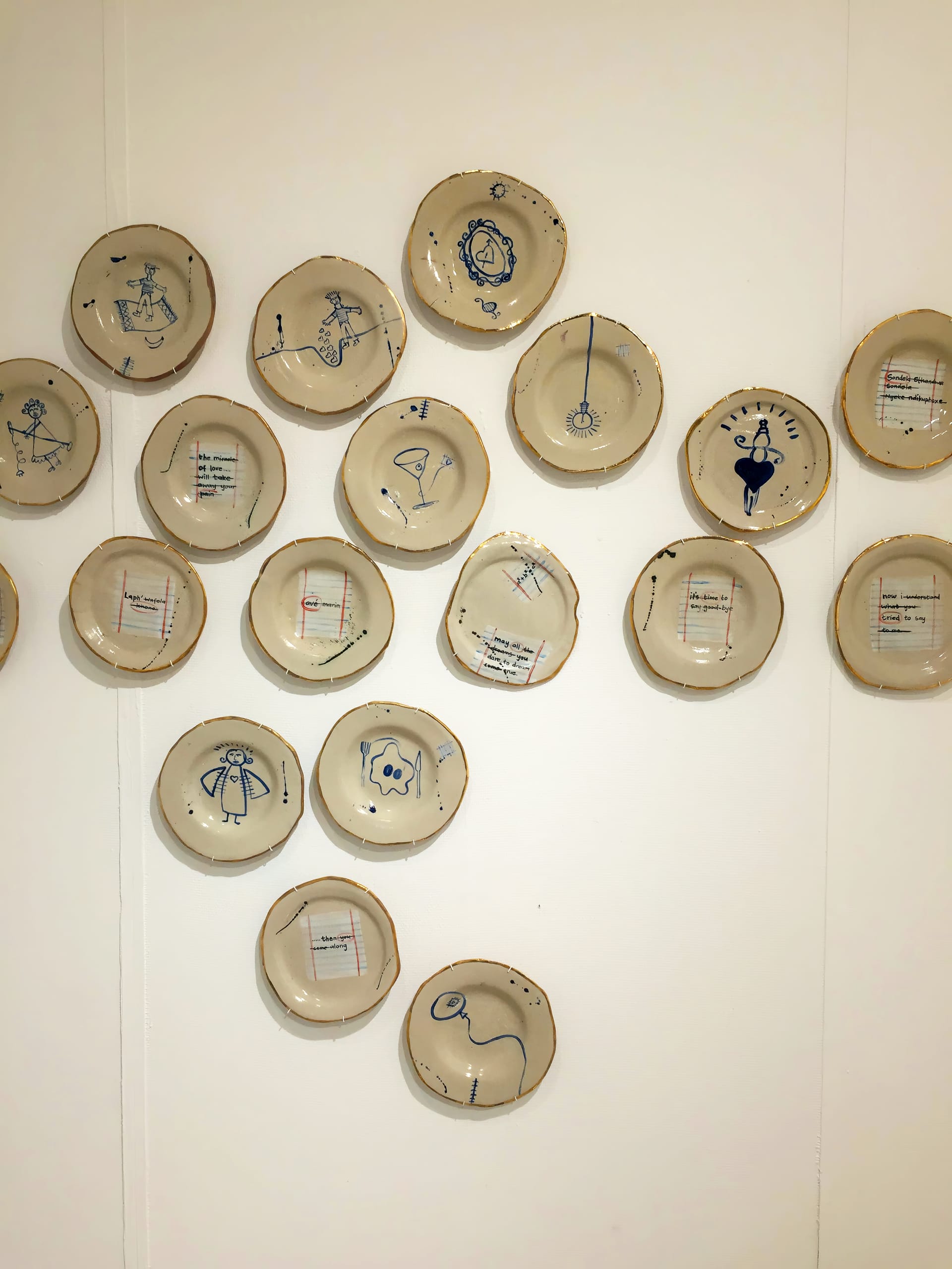 Twenty ceramic plates with drawings in blue are mounted on a white wall. The drawings depict simple, playful human or animal figures and objects. Some plates have handwritten text on lined paper, whilst others only have drawings. Plates are arranged loosely in a group.
